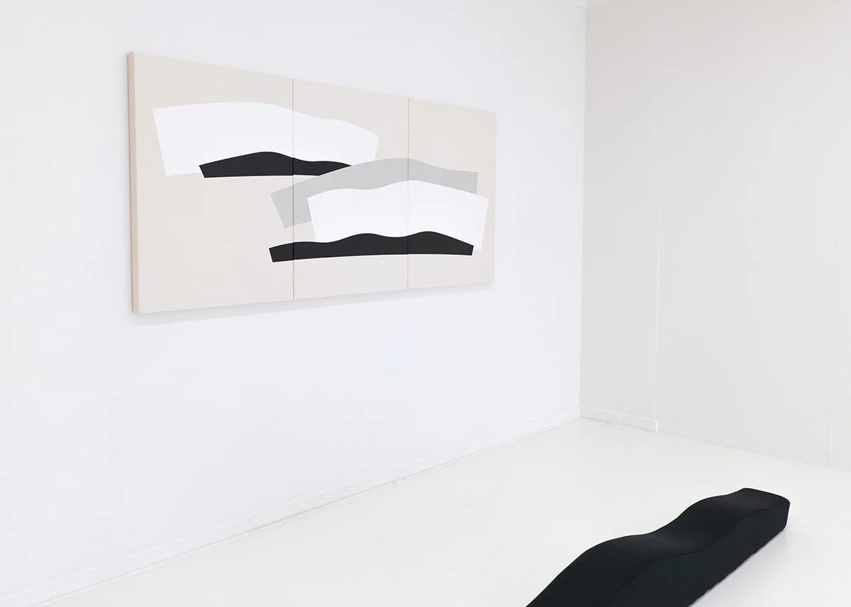 Minimalist art exhibit with a black bench on a white floor and a multi-panel abstract painting on the wall featuring black, white, and gray shapes.
