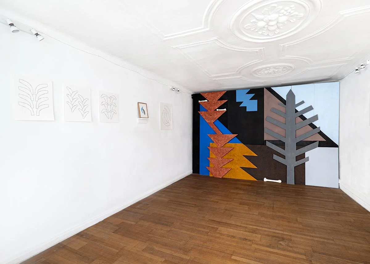 An art gallery featuring a white wall with framed white outlines of plants, and a large painting with abstract trees in bright colors on a corner wall.