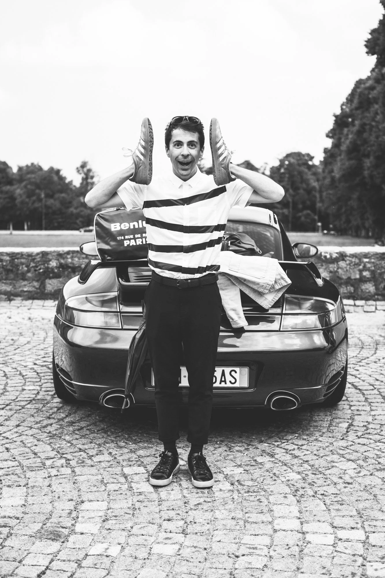 A young man standing in front of a Porsche 996 911 GT2, holding shoes to his ears, smiling, with luggage on the car's trunk, on a cobblestone pavement.
