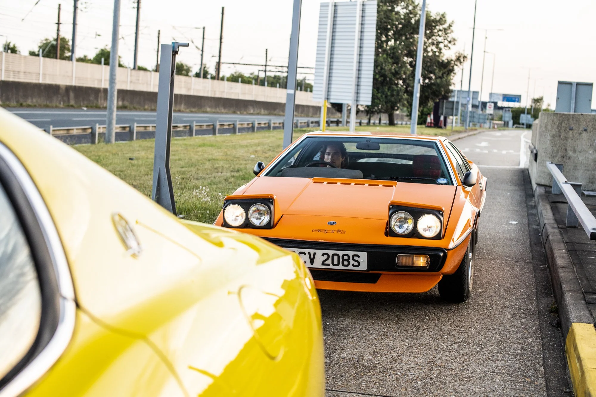 A Lotus Esprit car parked on the side of a road with a man inside, viewed through the windshield. Part of a yellow Maserati Quattroporte car is visible in the foreground.