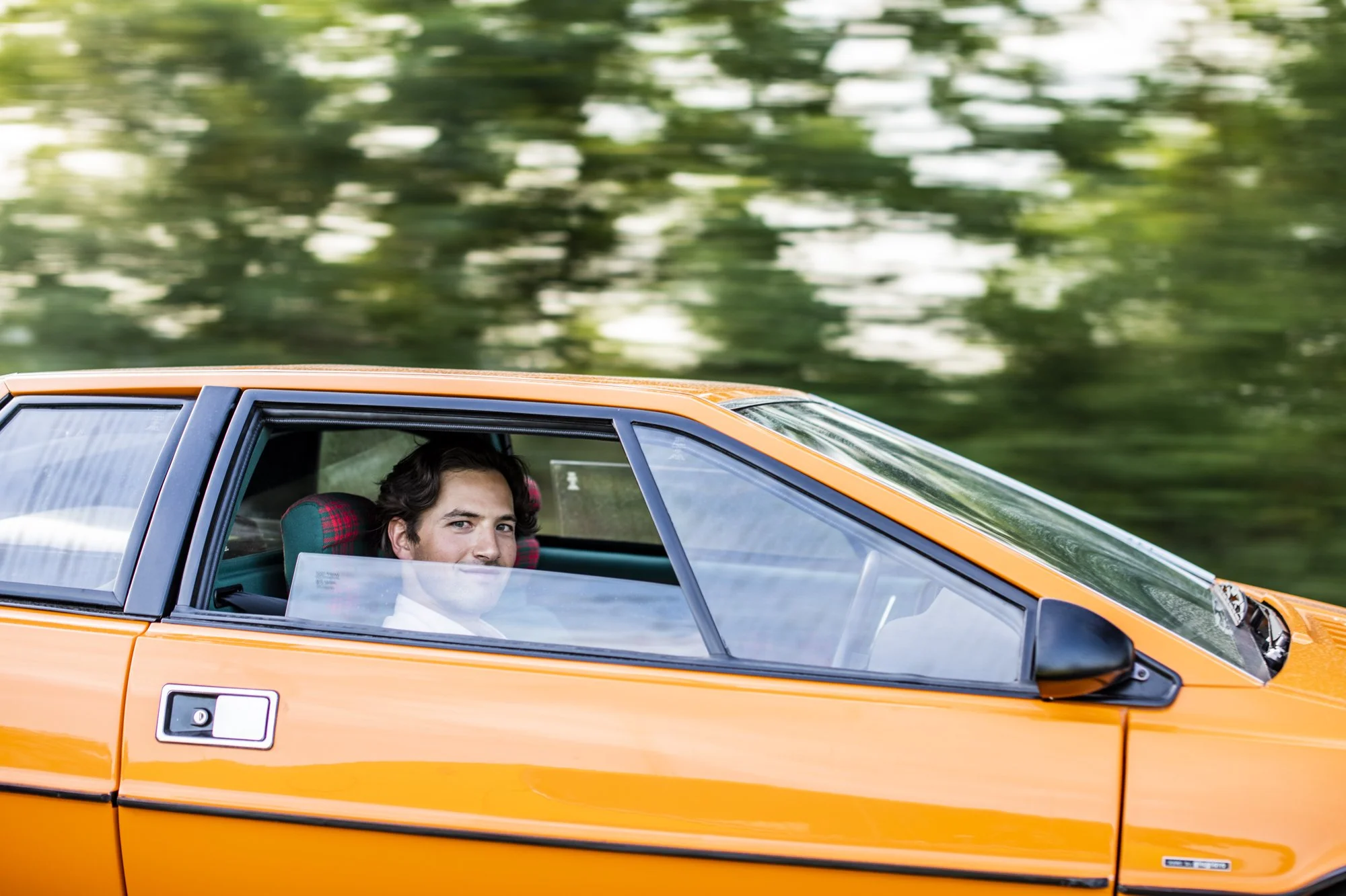 Young man with dark hair sitting in the driver's seat of Lotus Esprit, looking out the window with a smile, green blurred background.