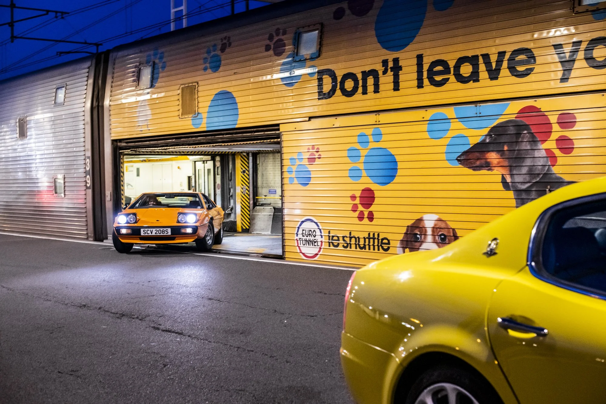 An Orange Lotus Esprit car driving out of a garage, with a yellow Maserati Quattroporte in the foreground, with a colorful wall mural and text that reads 'Don't leave you,' with a logo for 'Euro Tunnel' and 'leshuttle.'