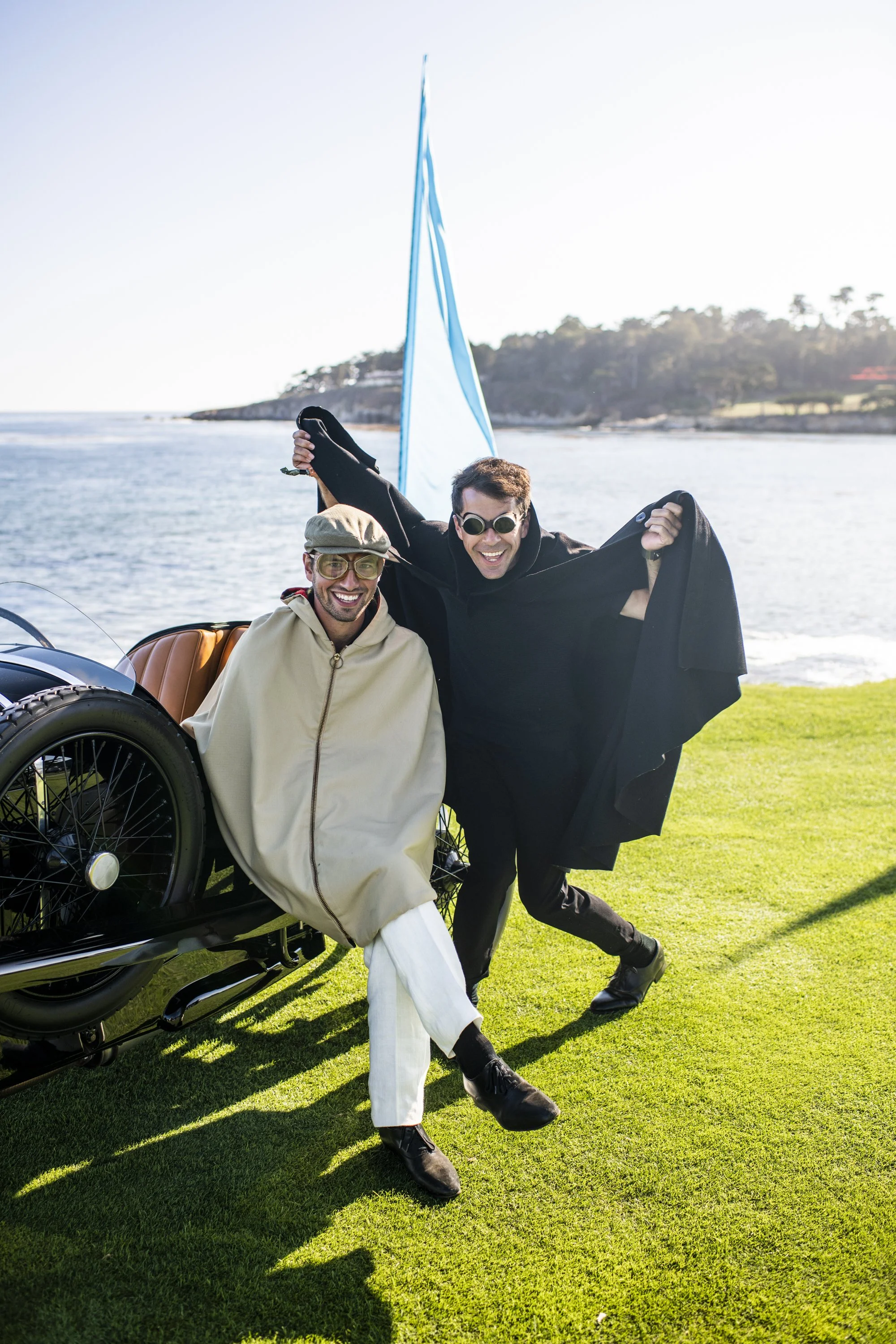 Two men dressed in trendy outfits are having fun by a shoreline with a vintage vehicle nearby. One man is sitting on the vehicle's side and the other is standing, both smiling and feeling joyful. The background shows water, a sailboat's sail, and distant trees and hills under a bright sky.