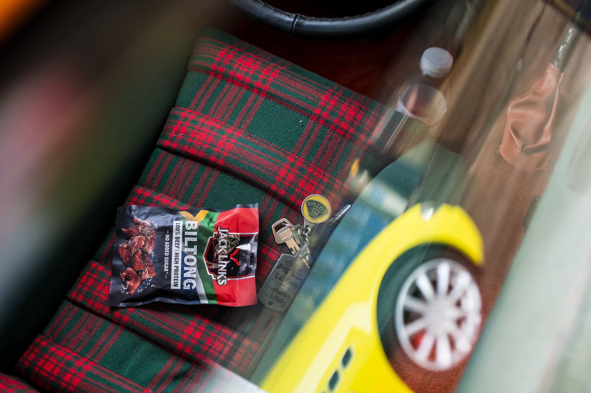 A yellow Maserati Quattroporte  with a black and silver wheel, a Jackslyn's Jack Links original beef jerky package, a set of keys with a keychain, a bottle of water, and a red and green plaid Lotus Esprit interior pattern.