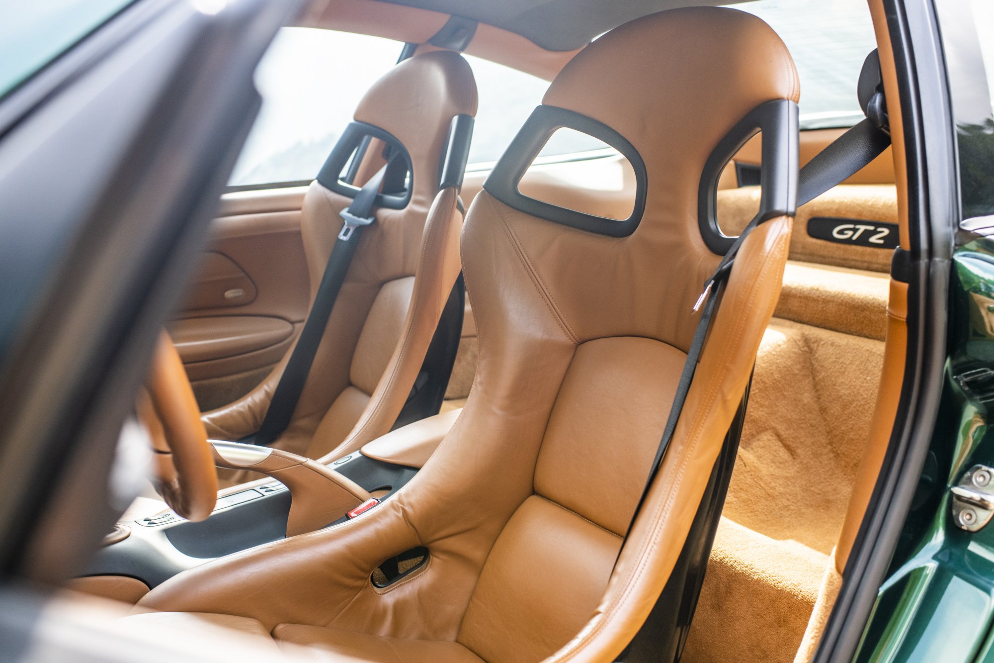 The interior of a Porsche 996 911 GT2 showing brown natural leather racing seats, black seat belts, and a tan carpeted rear area with a 'GT2' badge.