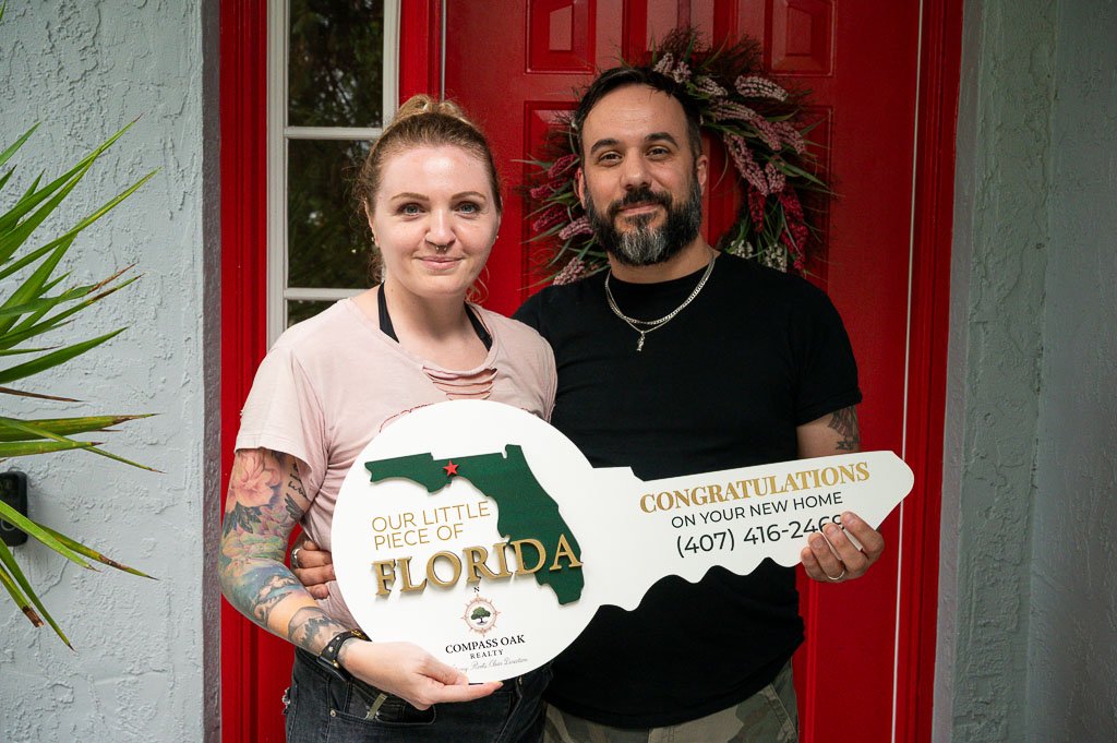 Happy Florida homebuyers standing in front of their new home holding a Compass Oak Realty congratulations key.