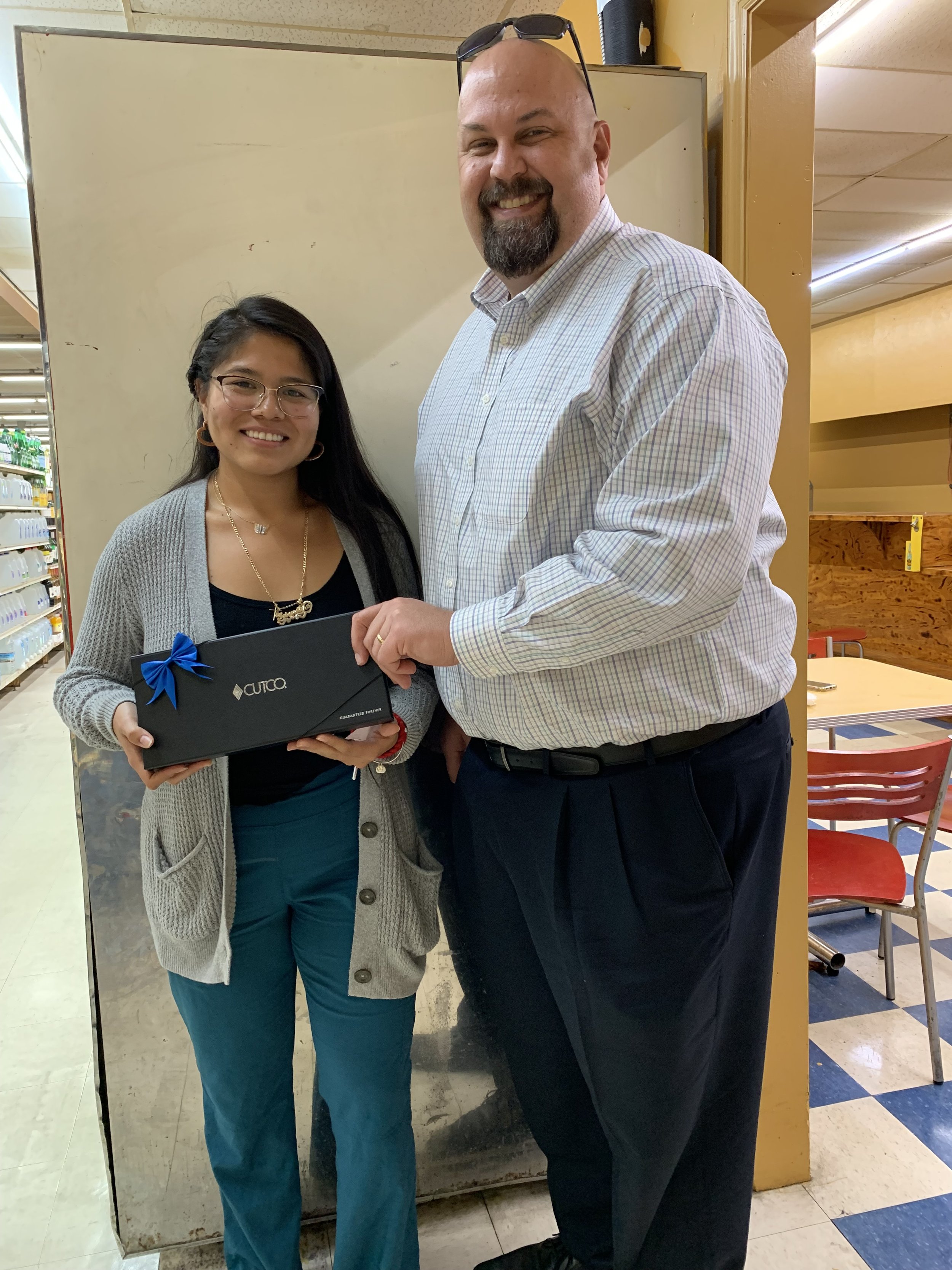 A young woman and an older man standing together indoors, smiling, with the woman holding a black gift box with a blue ribbon.
