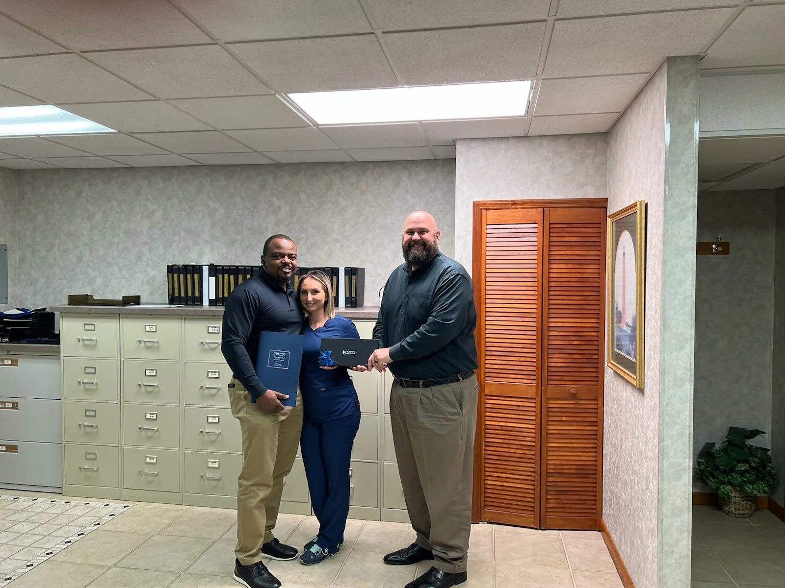 Three people standing indoors, two men and one woman, exchanging a gift and a folder, smiling, in an office setting with filing cabinets and framed picture