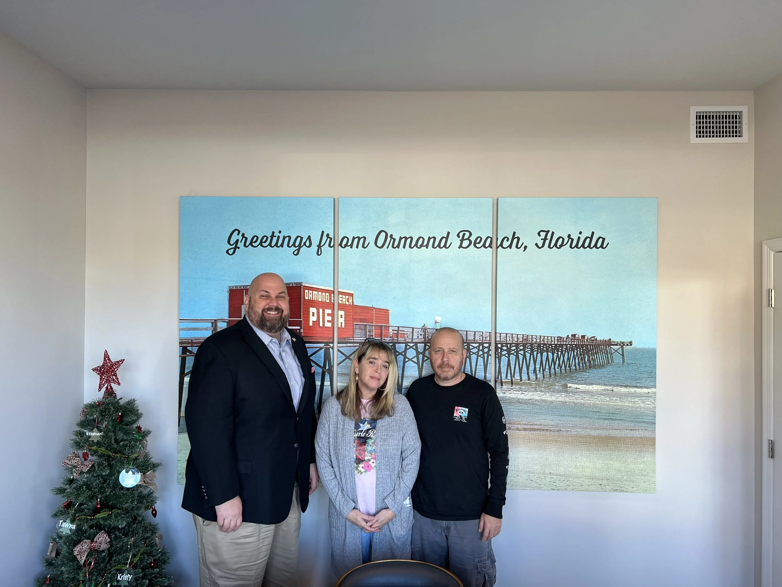 Three people standing in front of a large wall art that says "Greetings from Ormond Beach, Florida" and shows a pier extending into the ocean; a decorated Christmas tree is to the left.