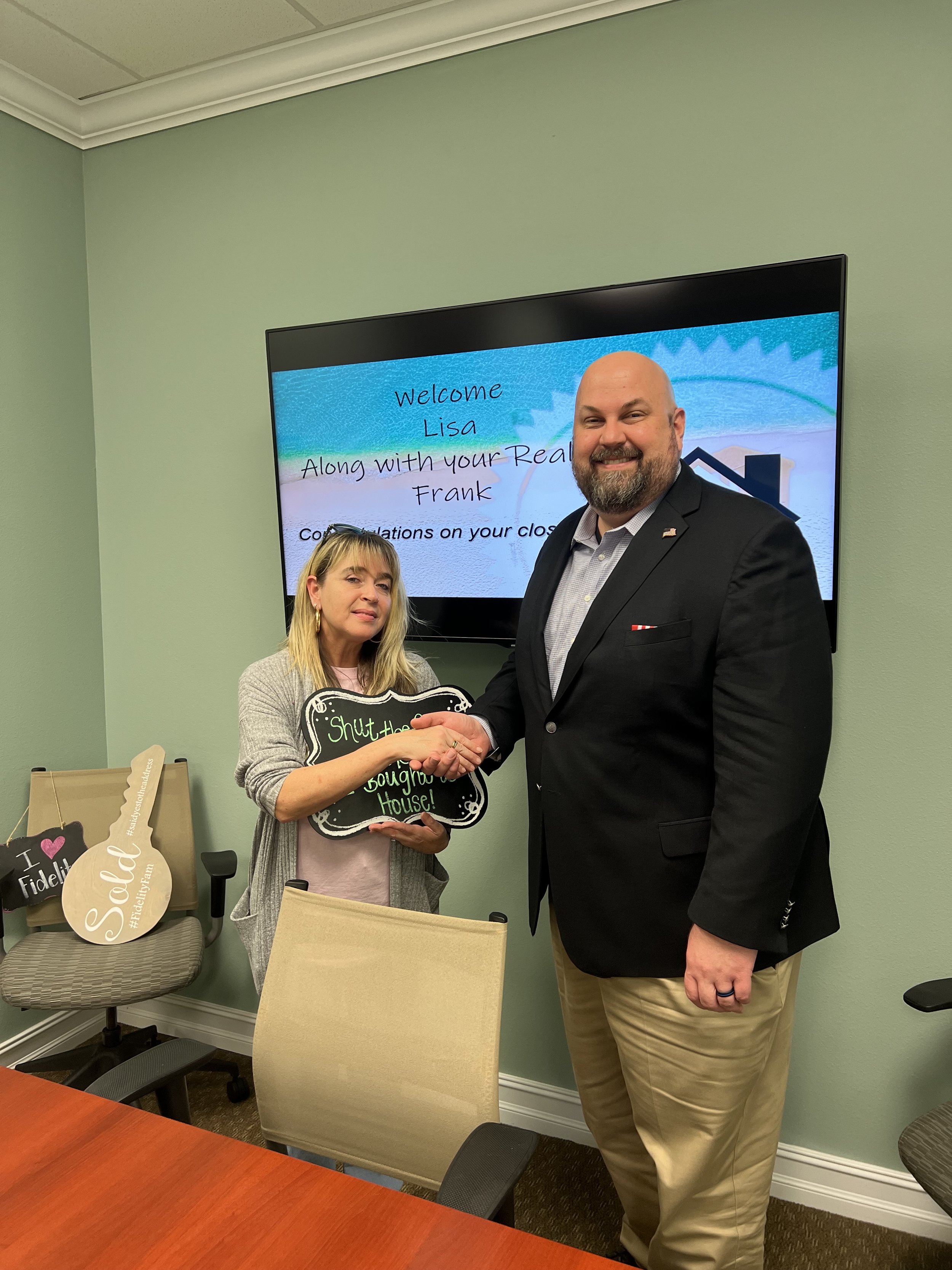 A man and woman shaking hands in a room with a green wall. The woman holds a speech bubble sign that says 'Shut the 'Do Not Disturb' House!'. Behind them, a large screen displays a welcome message for Lisa and Frank, congratulating on their closing.