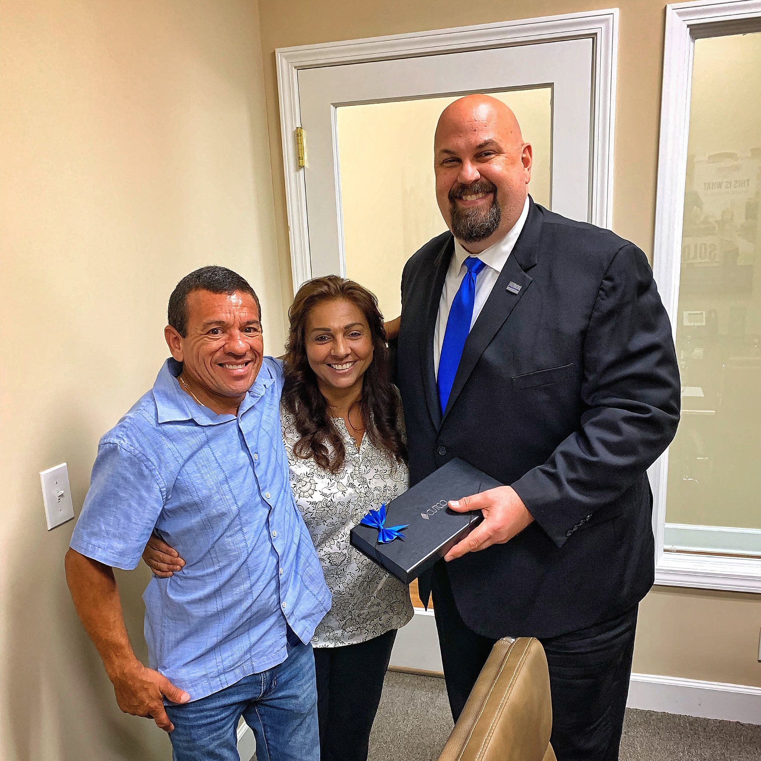 Three people standing inside an office, smiling at the camera. The man on the right is holding a box with a blue ribbon.