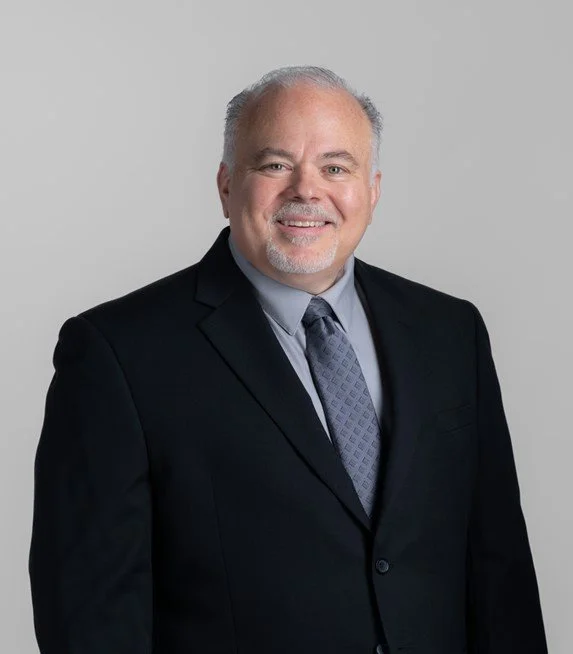 A middle-aged man in a black suit, light blue shirt, and patterned tie, smiling against a plain gray background.