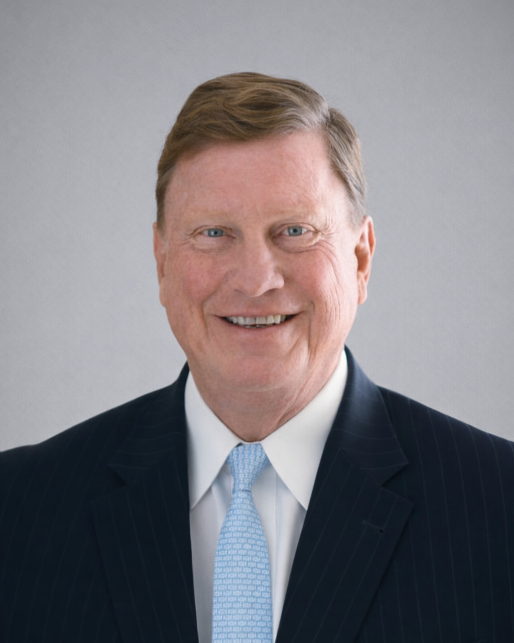 Tom Fanning, a man with blue eyes, light brown hair, wearing a dark pinstripe suit, white shirt, and light blue tie, against a plain gray background.