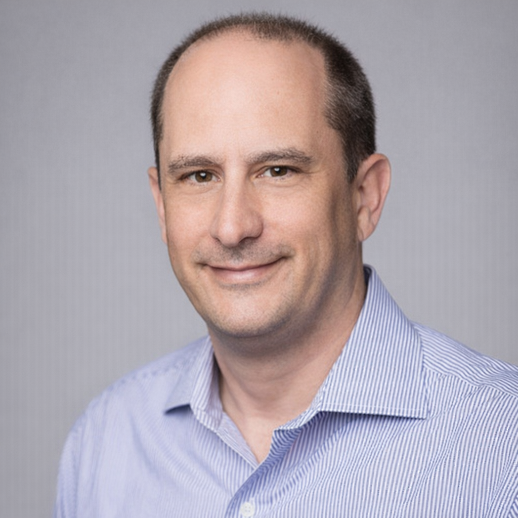 Portrait of a man with short brown hair, light skin, wearing a light blue collared shirt, smiling against a gray background.