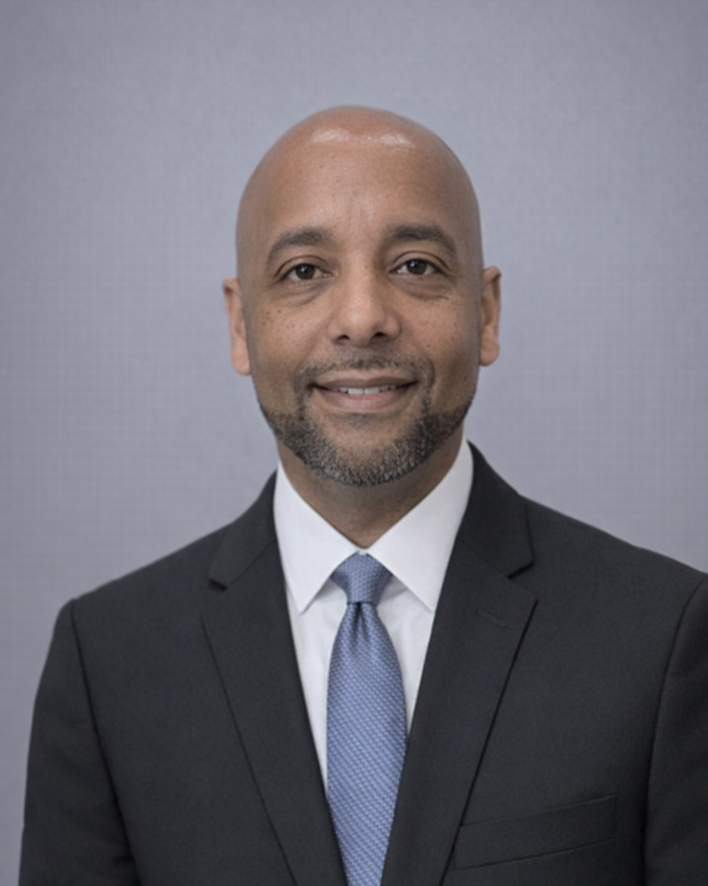 Professional headshot of a man in a black suit, white shirt, and light blue tie, smiling against a gray background.