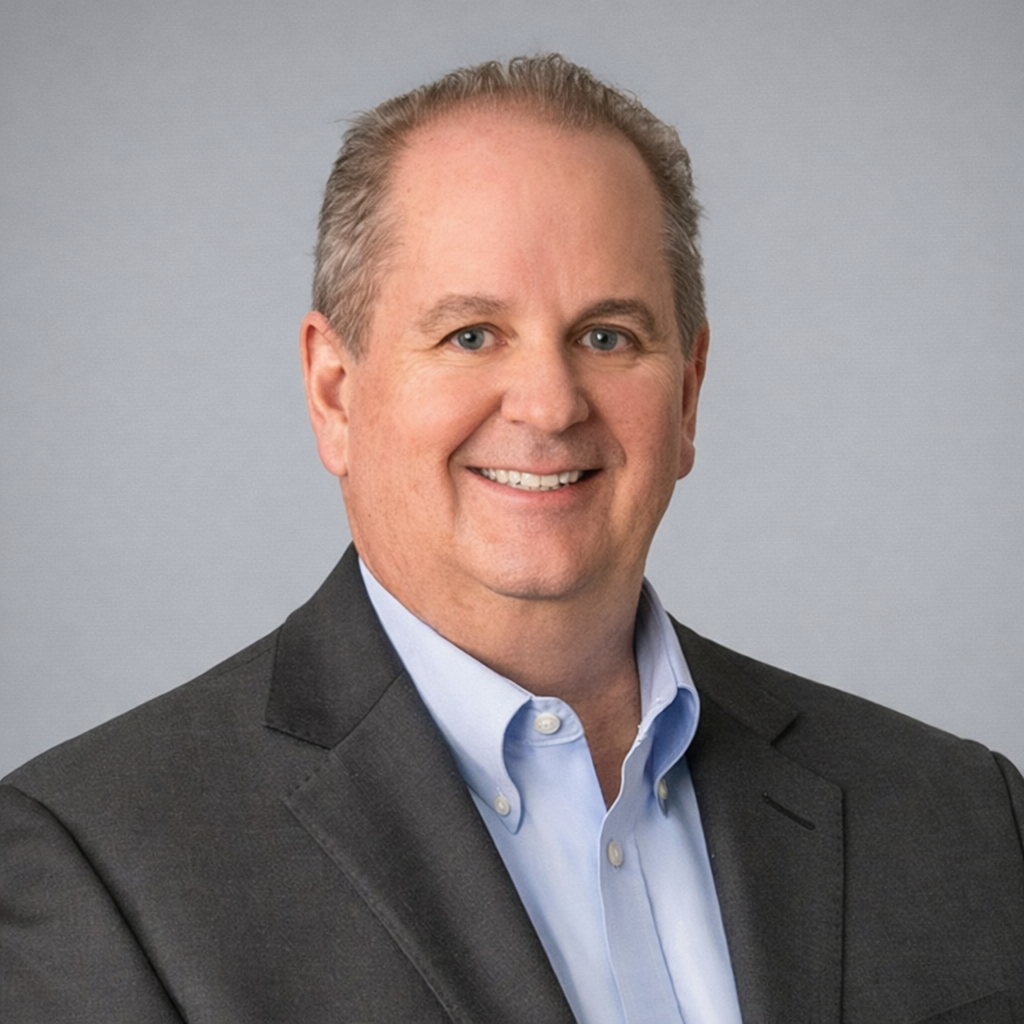 A professional headshot of a middle-aged man with short, gray hair, wearing a dark suit jacket and a light blue collared shirt, smiling against a light gray background.