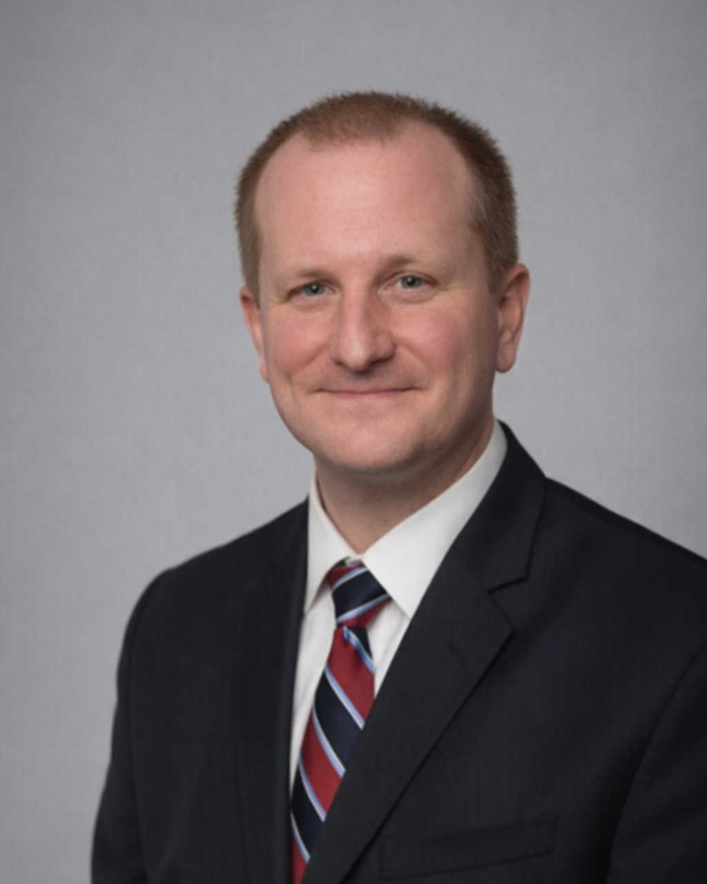 Jeff Baumgartner, a man wearing a suit and tie, smiling, against a plain gray background.