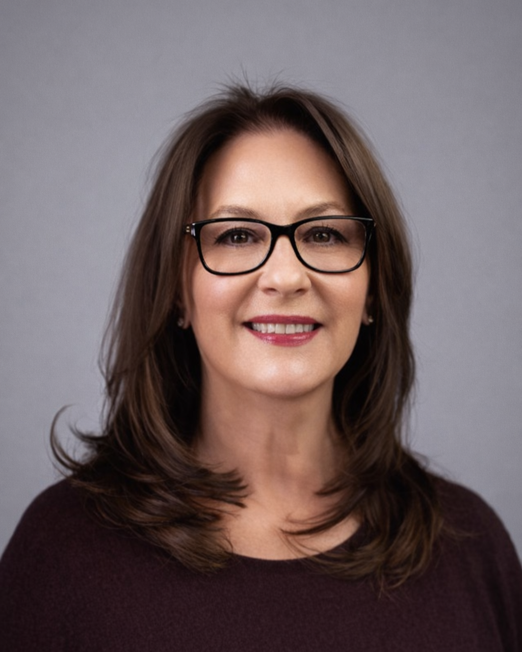 Michele Guido, a woman with medium-length brown hair, black glasses, and a dark top, smiling in front of a plain gray background.