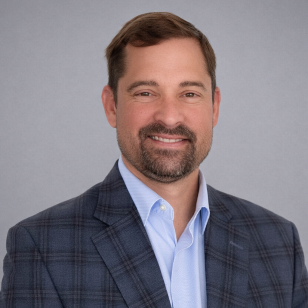 Headshot of a man with brown hair, a beard, wearing a blue dress shirt and a plaid blazer, smiling against a gray background.