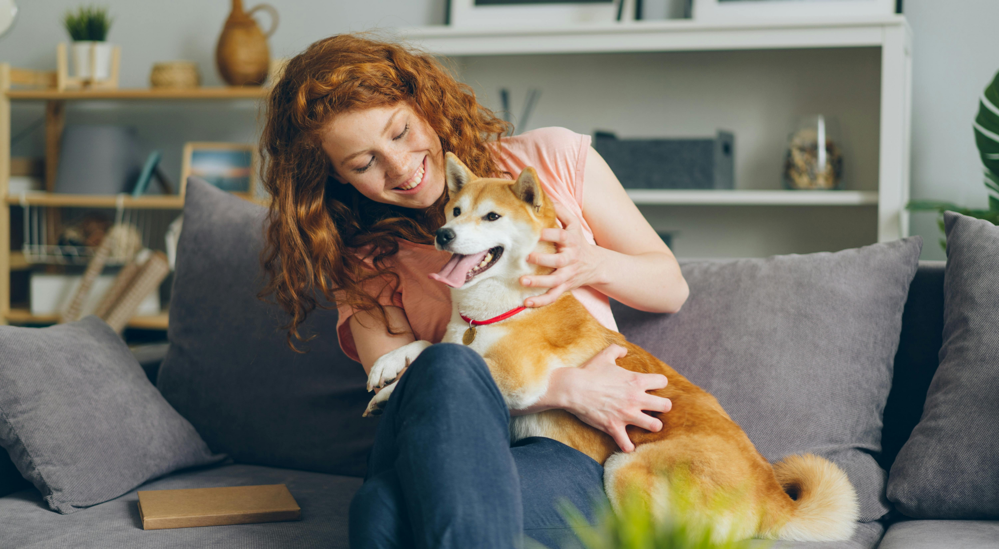 Una mujer con cabello rizado color rojo, sonriendo, mientras abraza a un perro de raza Shiba Inu en un sofá gris en un ambiente hogareño.