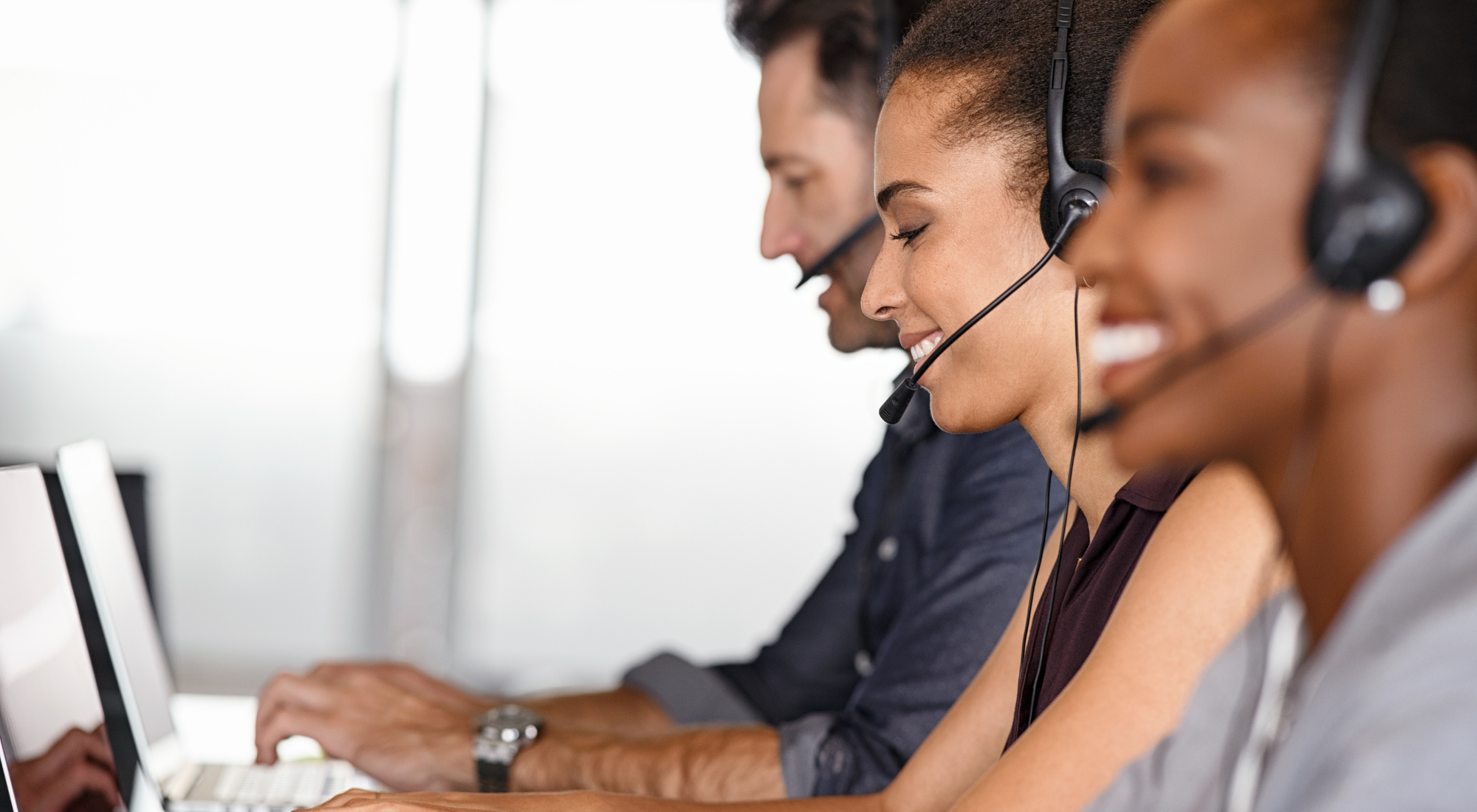 Tres personas con auriculares de oficina trabajando en computadoras en fila.