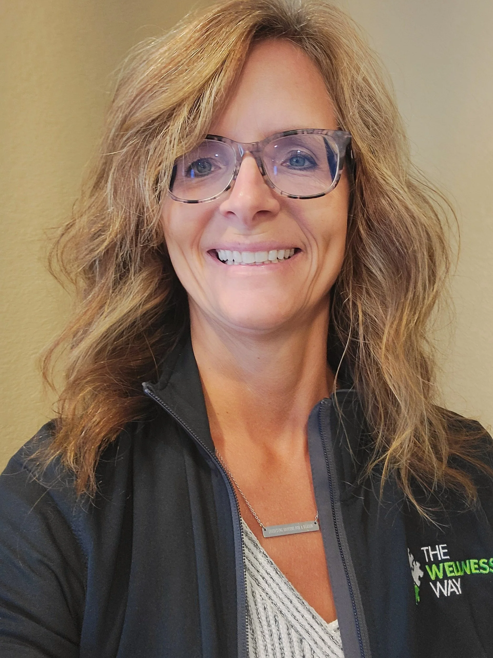 A woman with curly blonde hair wearing glasses, a black jacket with a logo that says "The WEINESS WAY," and a silver necklace smiling at the camera against a beige wall.