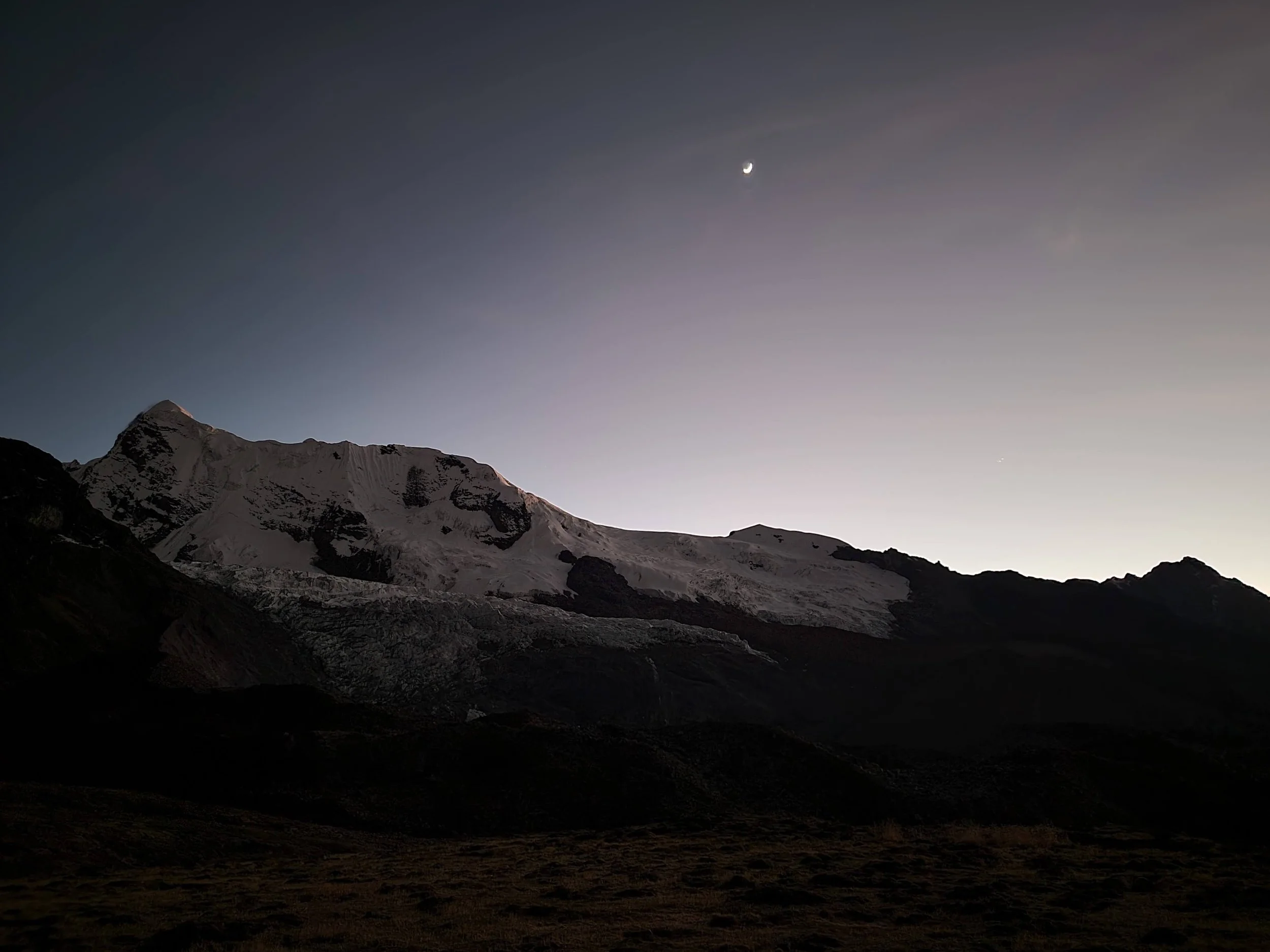 Snow-covered mountain peaks at dusk or dawn with visible crescent moon and a bright celestial object in the sky.