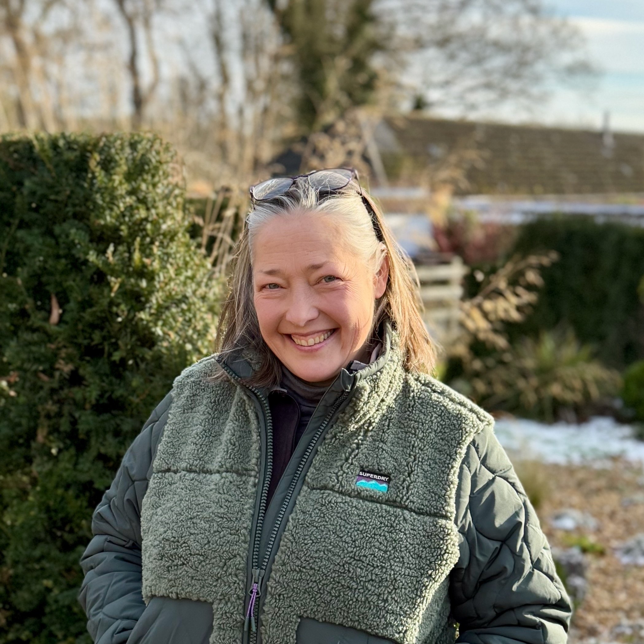 Smiling woman outdoors in a green fleece and jacket, with glasses on her head, autumn trees, and a house in the background.