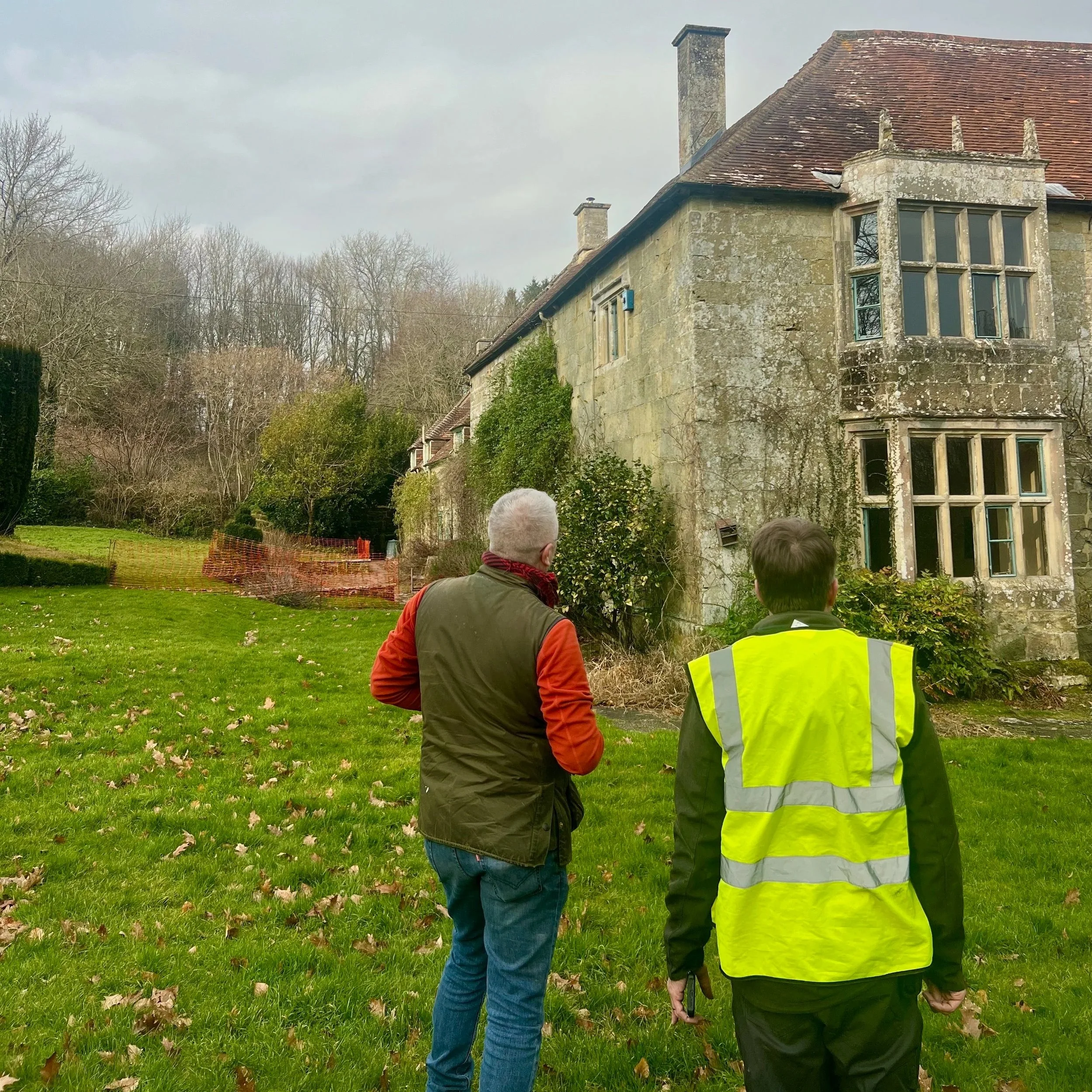 Two men, one with gray hair wearing a green vest over an orange jacket and the other with brown hair in a yellow high-visibility vest, standing on a lawn and looking at an old, weathered stone house with overgrown vegetation and broken windows.