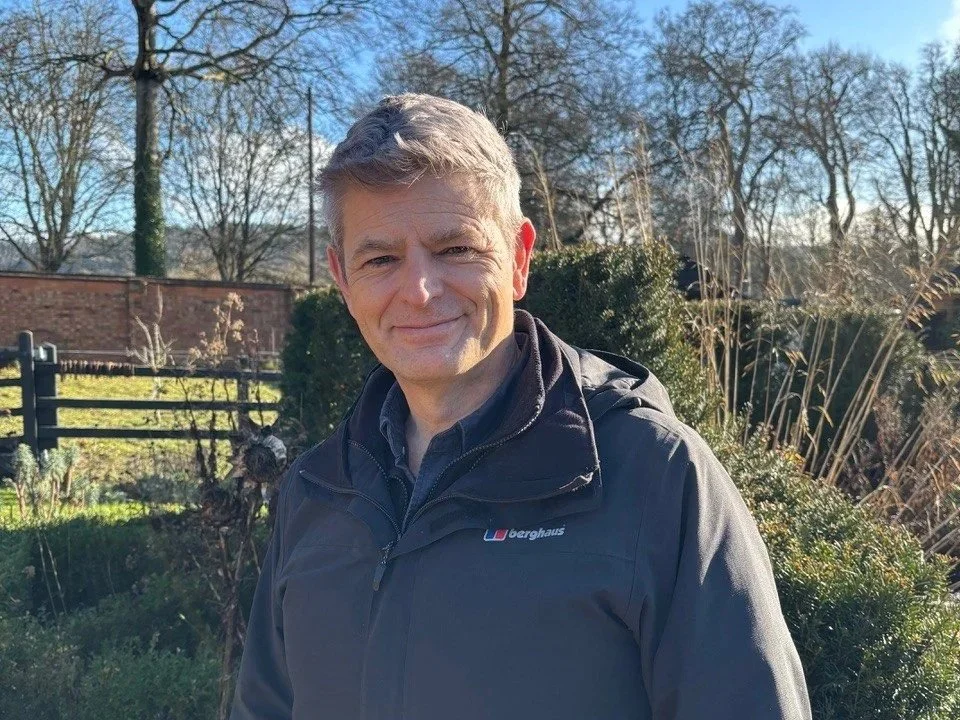 A man outdoors smiling, wearing a black jacket, with a yard and bare trees in the background.