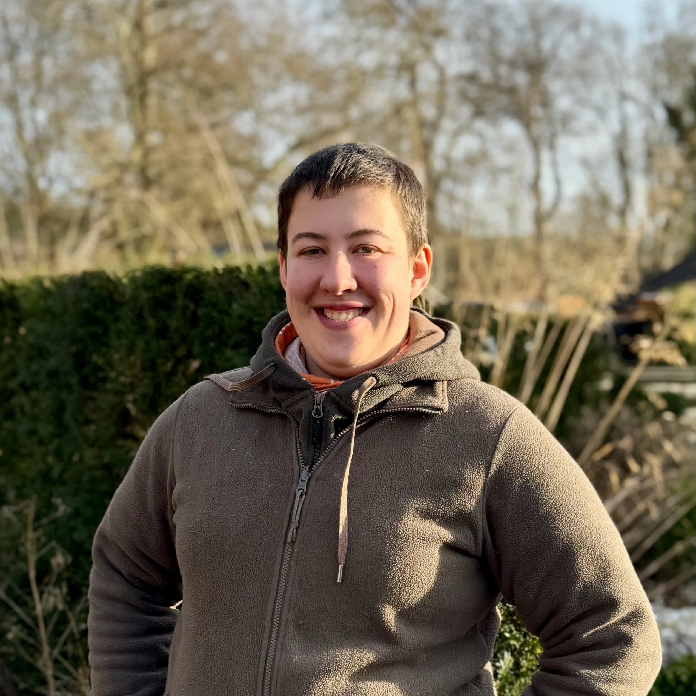 A young person smiling outdoors in a cozy brown zip-up hoodie with a hood, standing in front of a hedge and leafless trees during daytime.