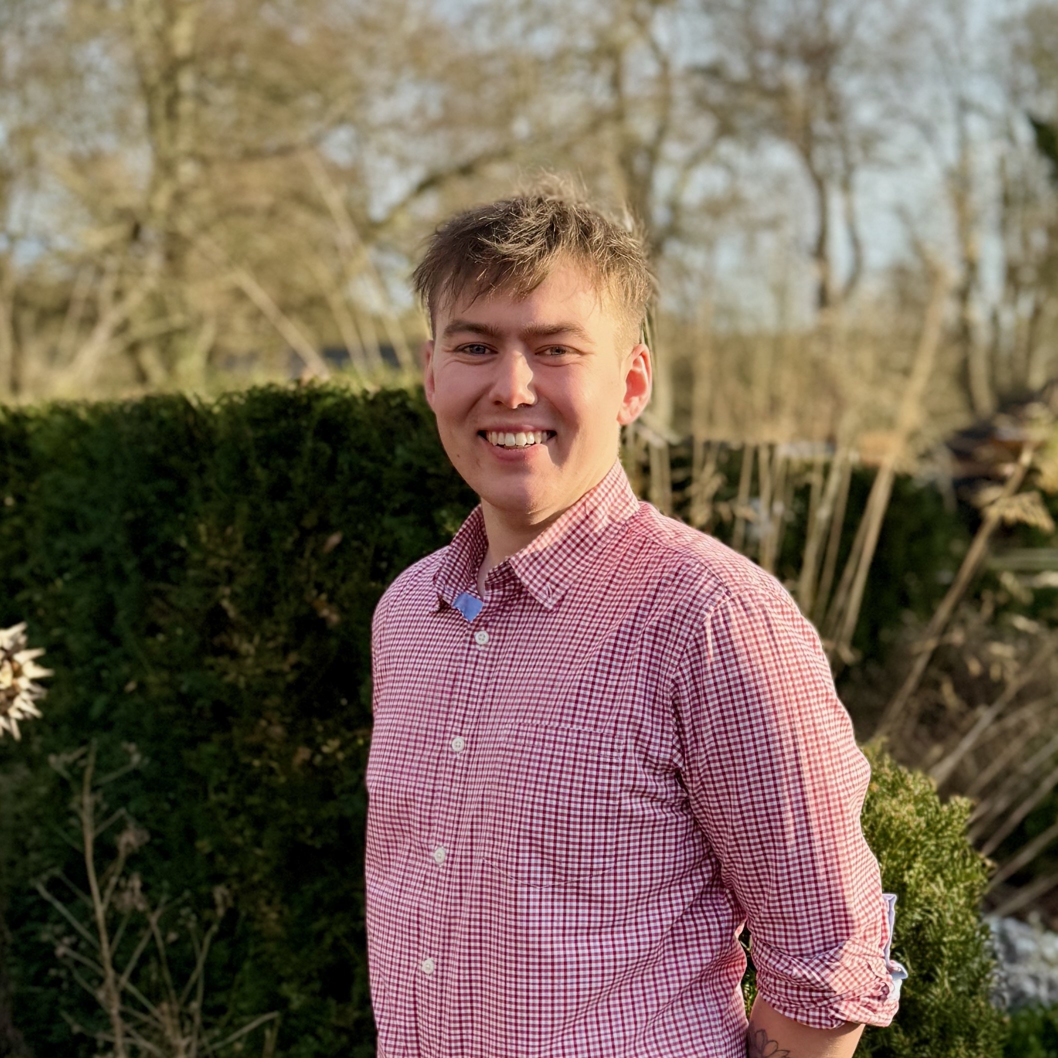 A young man with short light brown hair and light skin, smiling outdoors during daylight, wearing a red and white checkered button-up shirt, standing in front of a green bush and leafless trees.