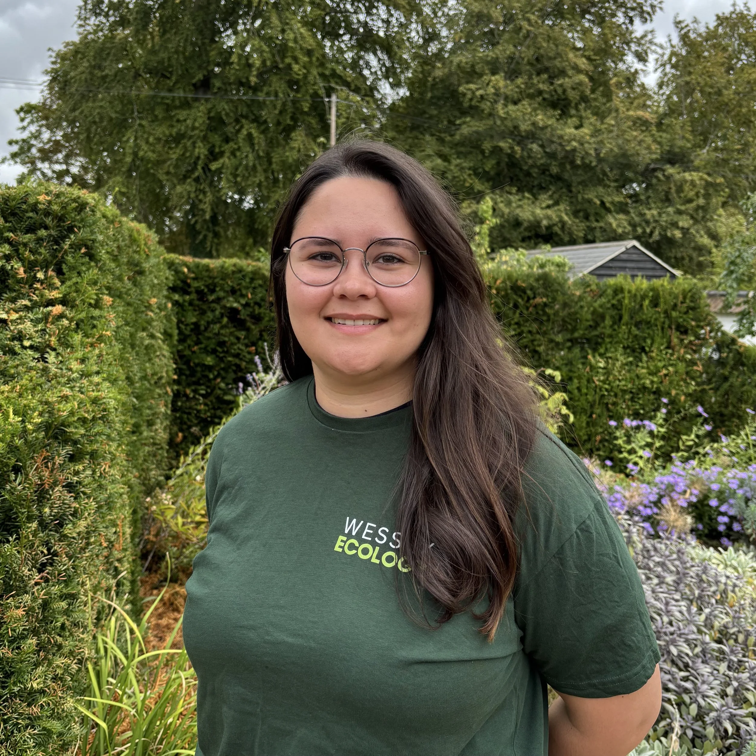 A young woman with long dark hair smiling outdoors in front of bushes and trees during sunset.