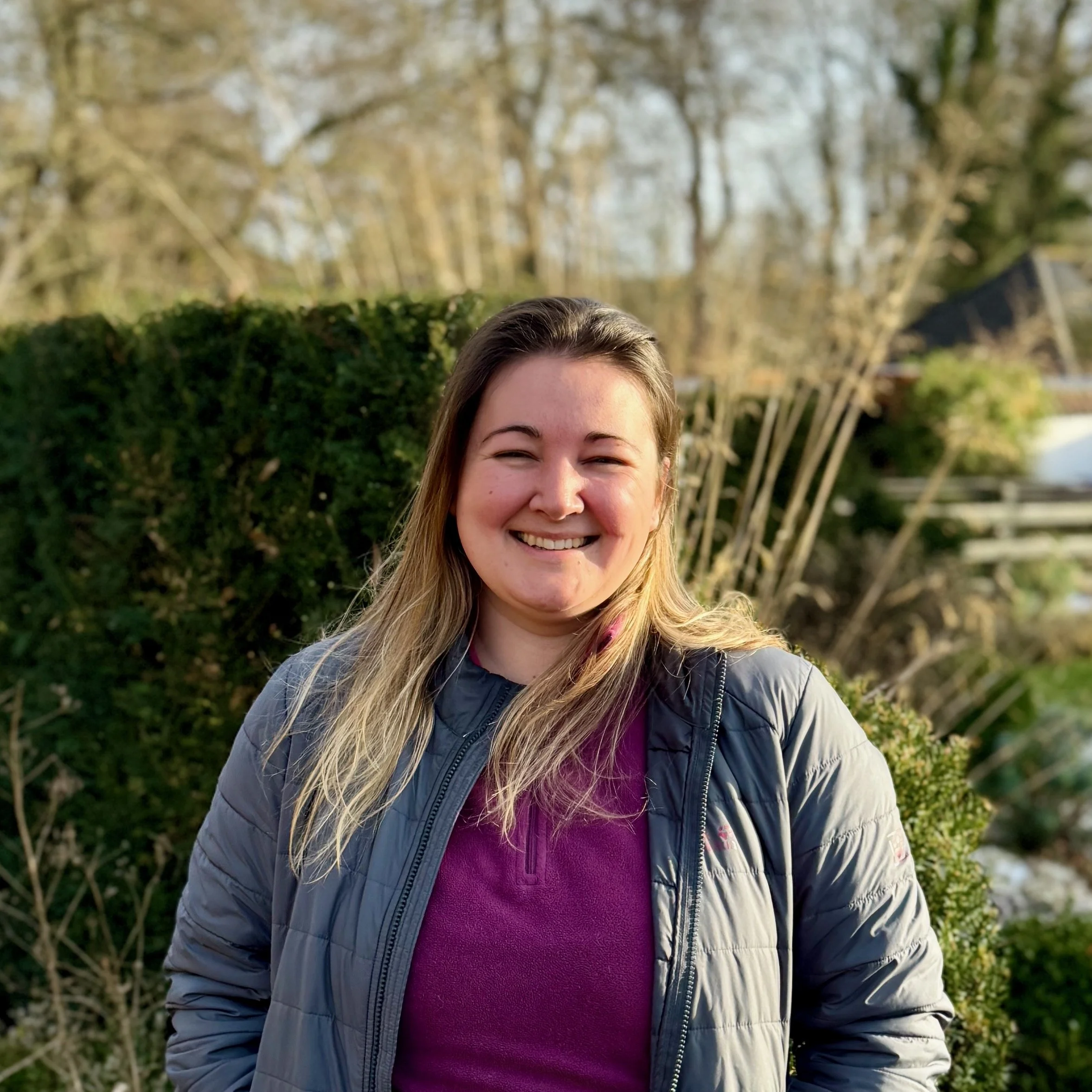 A woman smiling outdoors with trees and greenery in the background.