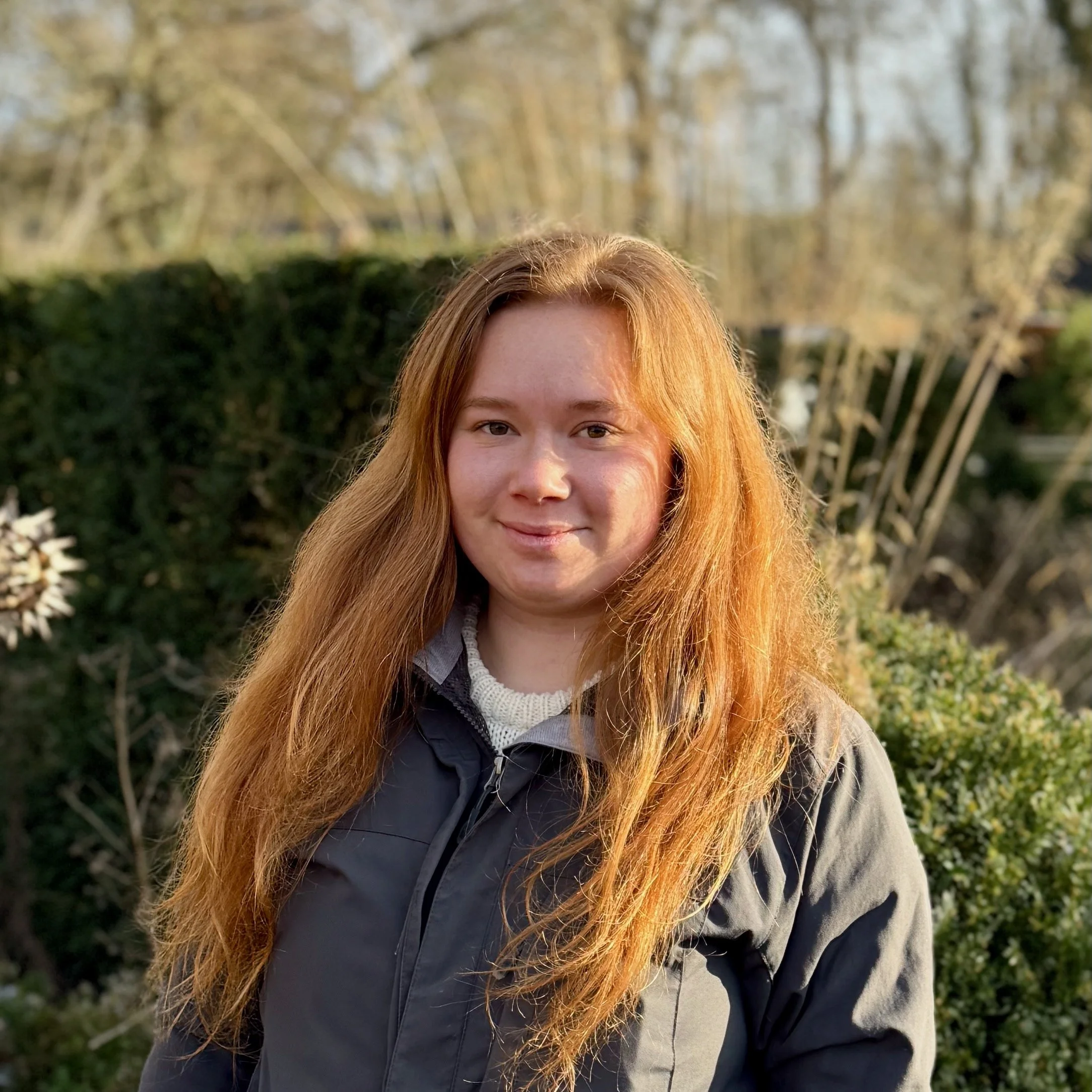 A young woman with long red hair smiling outdoors in front of bushes and trees during sunset.