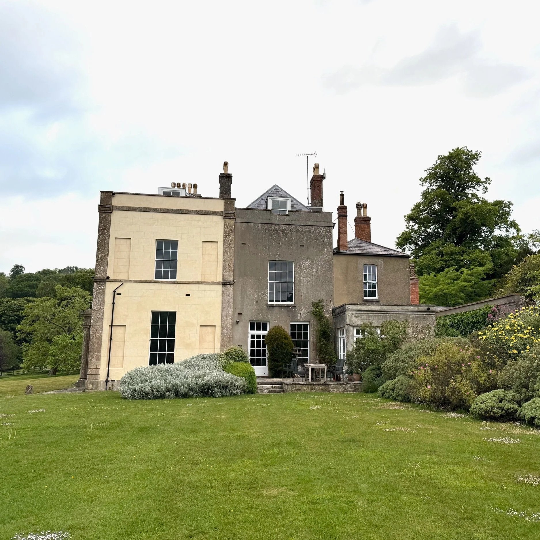A historic multi-story house with a mix of stone and brick exterior, multiple chimneys, and large windows, surrounded by a well-maintained lawn and garden with bushes and trees under a cloudy sky.