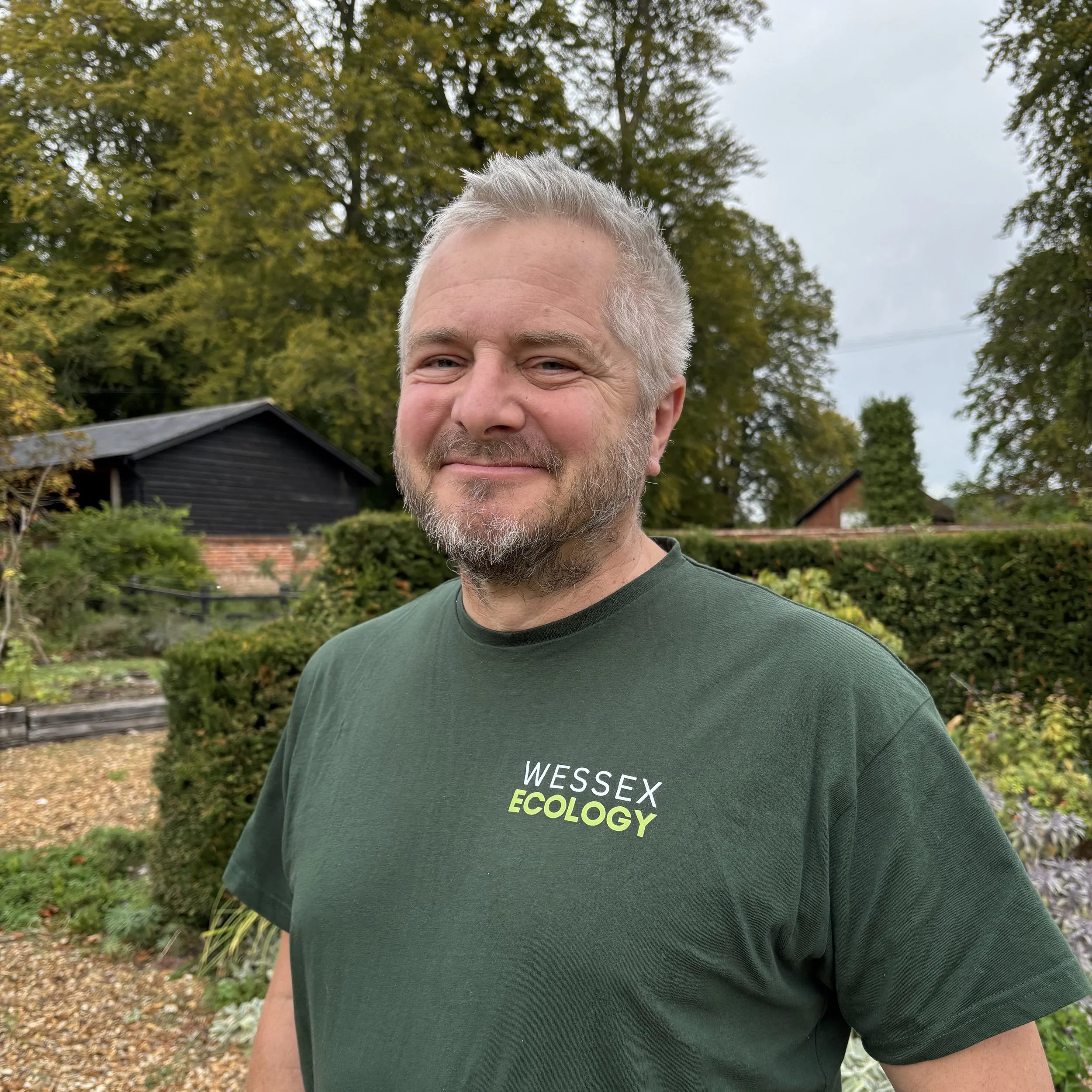 A man with grey hair and a beard smiling outdoors in a garden with trees and fences in the background, wearing a green jacket.