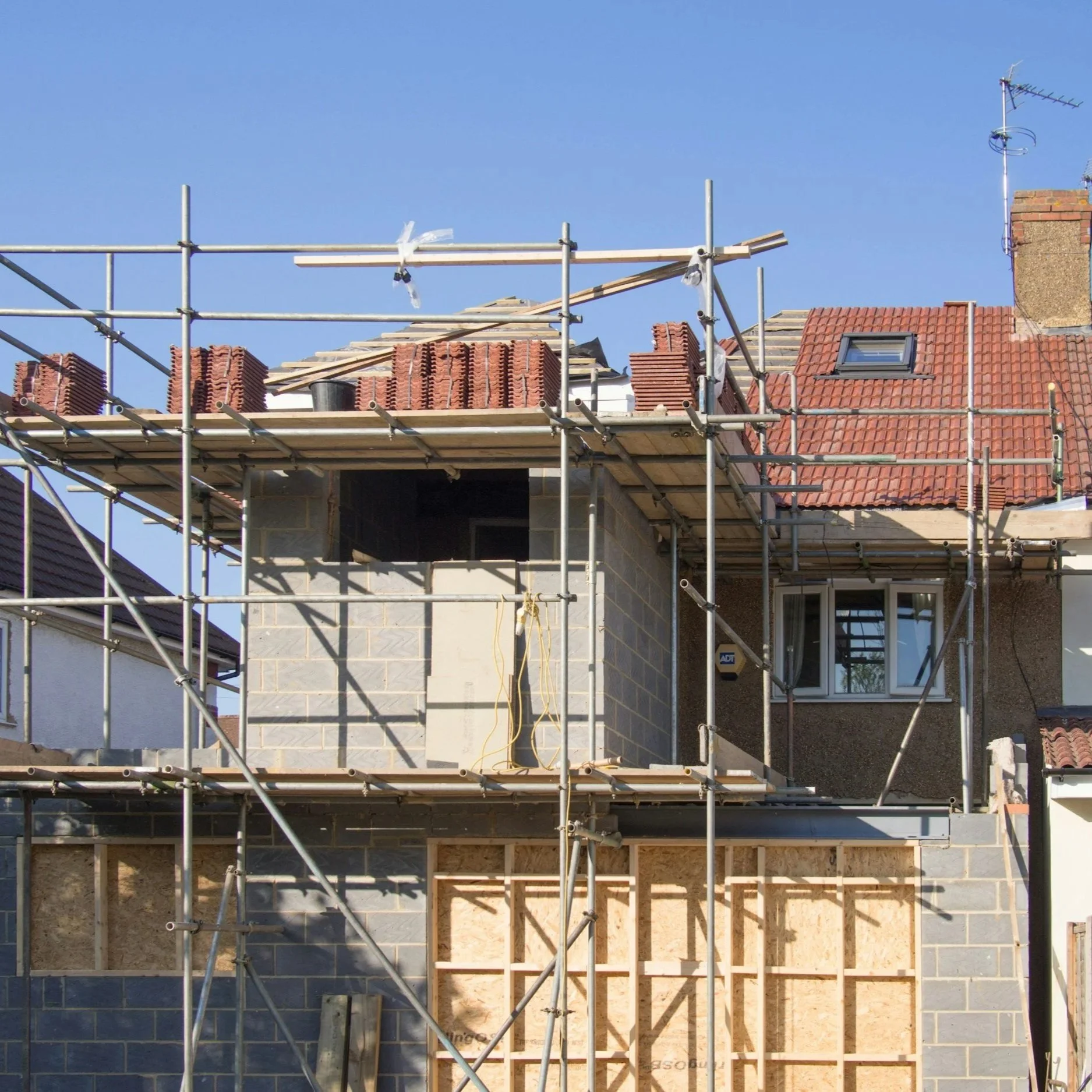 Construction site of a building with scaffolding, partially built concrete walls, and a roof under construction with stacks of bricks and wooden planks under a clear blue sky.
