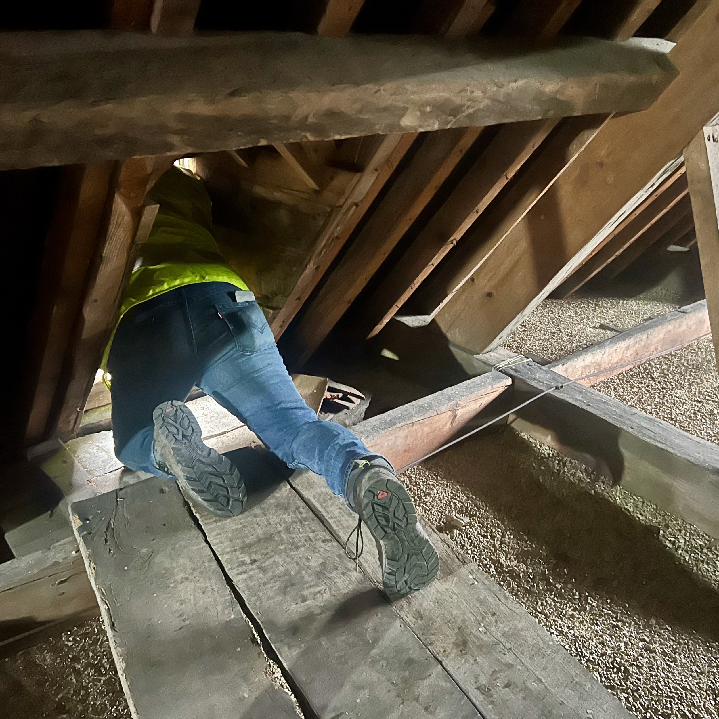 Worker crouching underneath a wooden structure, inspecting or working on foundation work in a construction site, wearing a yellow safety vest and jeans.