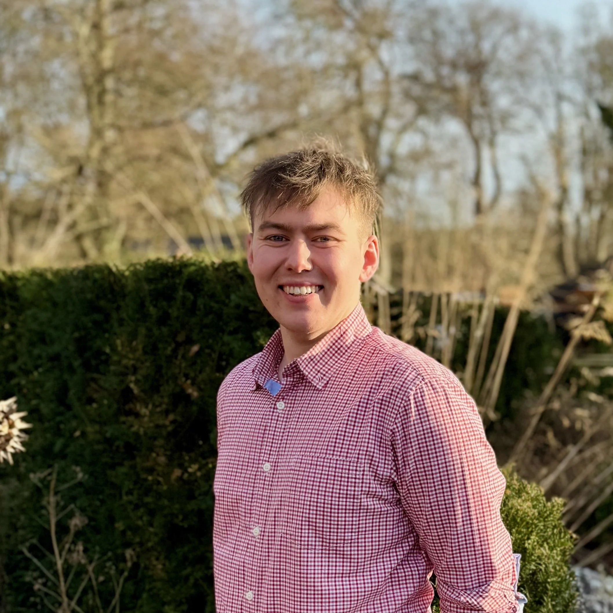 A young man with short light brown hair and light skin, smiling outdoors during daylight, wearing a red and white checkered button-up shirt, standing in front of a green bush and leafless trees.