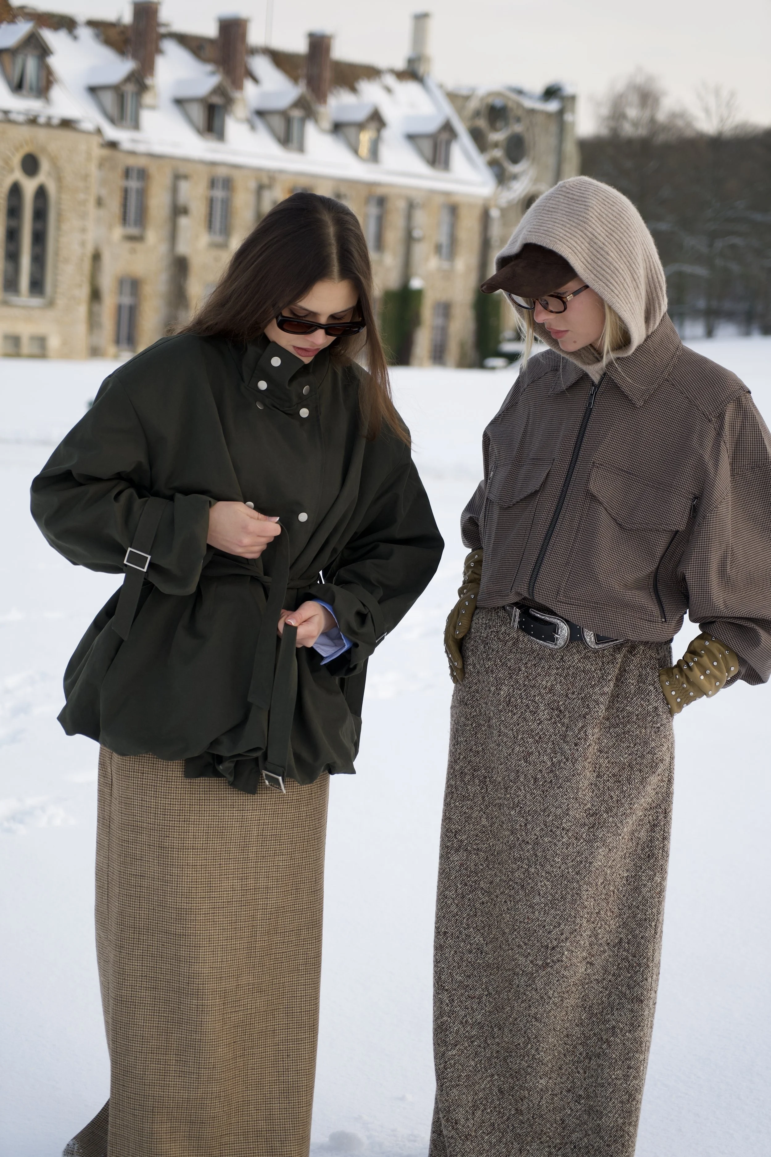 Deux femmes habillées en tenues d'hiver élégantes se tiennent dans un paysage enneigé devant une abbaye ancienne.