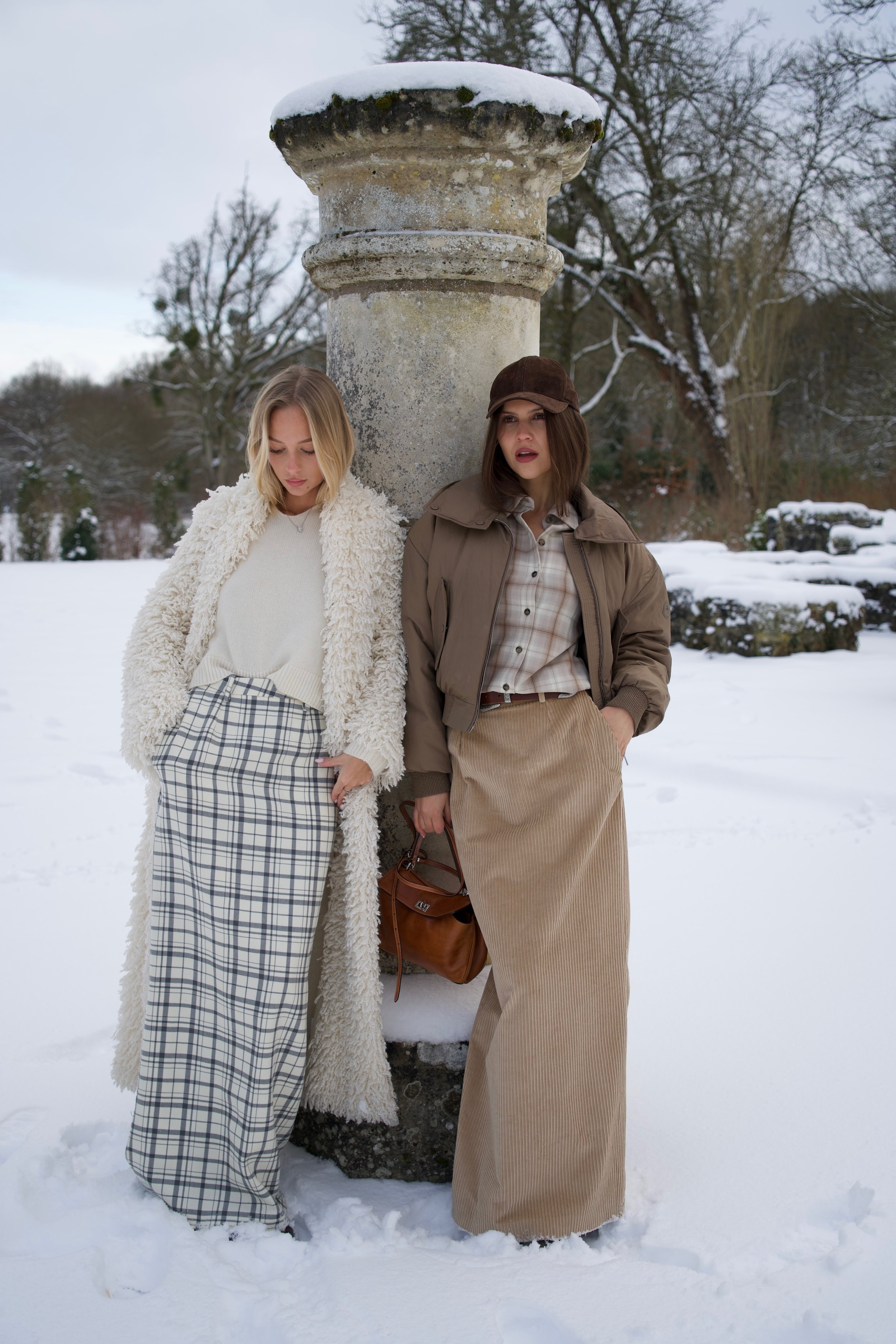 Deux femmes posant dans un paysage enneigé, vêtues de vêtements d'hiver tendance, en dehors d'une ancienne colonne en pierre.
