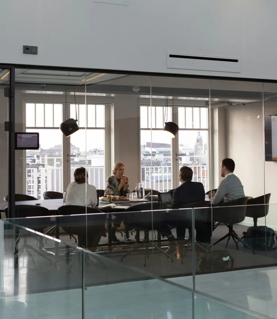 Cuatro personas en una sala de reuniones con ventanas grandes, dos mujeres y dos hombres sentados en una mesa, participando en una discusión o reunión de trabajo.