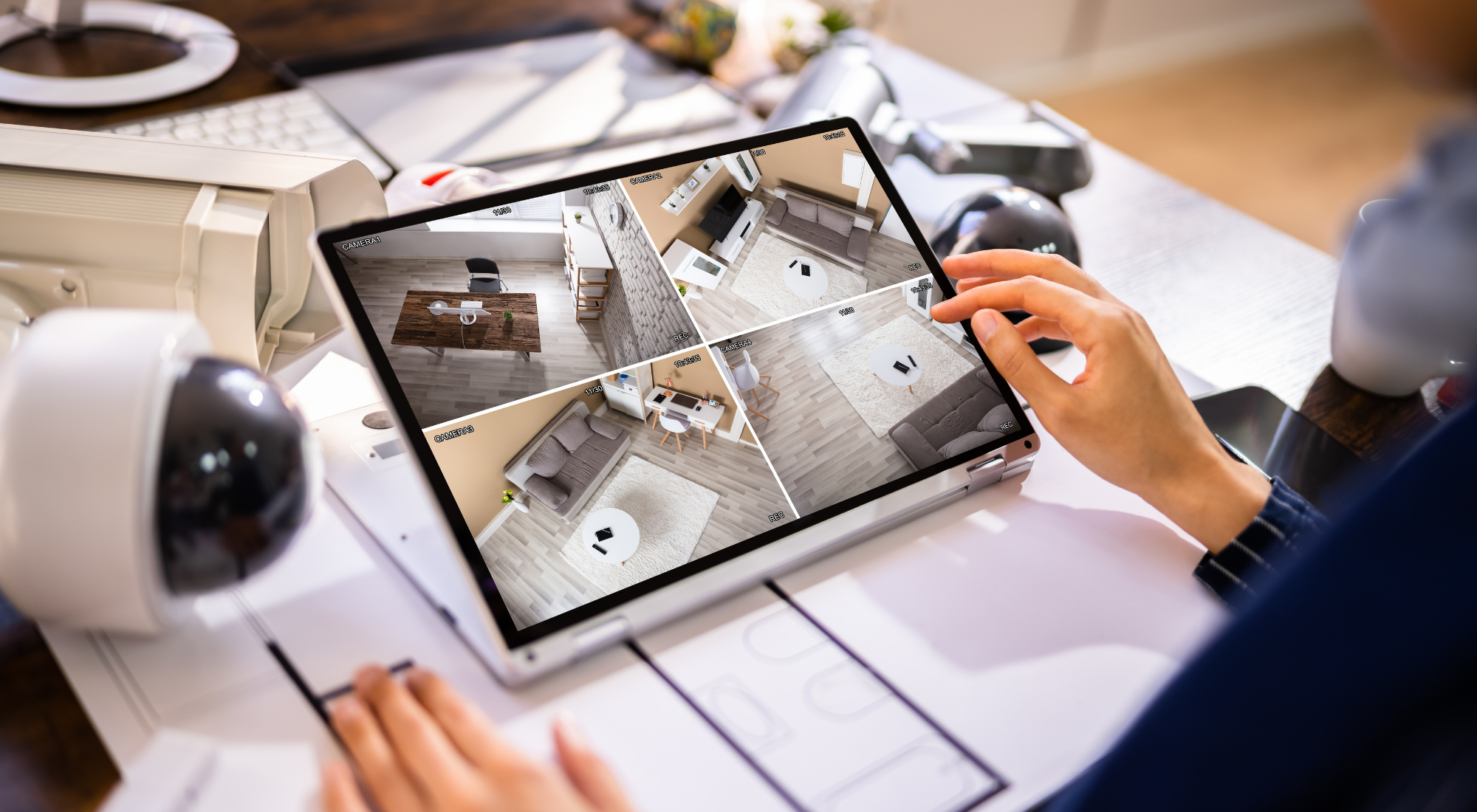 Una mano tocando una tableta que muestra una vista en vivo de varias cámaras de seguridad en diferentes habitaciones de una casa, con dispositivos electrónicos como una videocalculadora, una cámara y una impresora en la mesa.