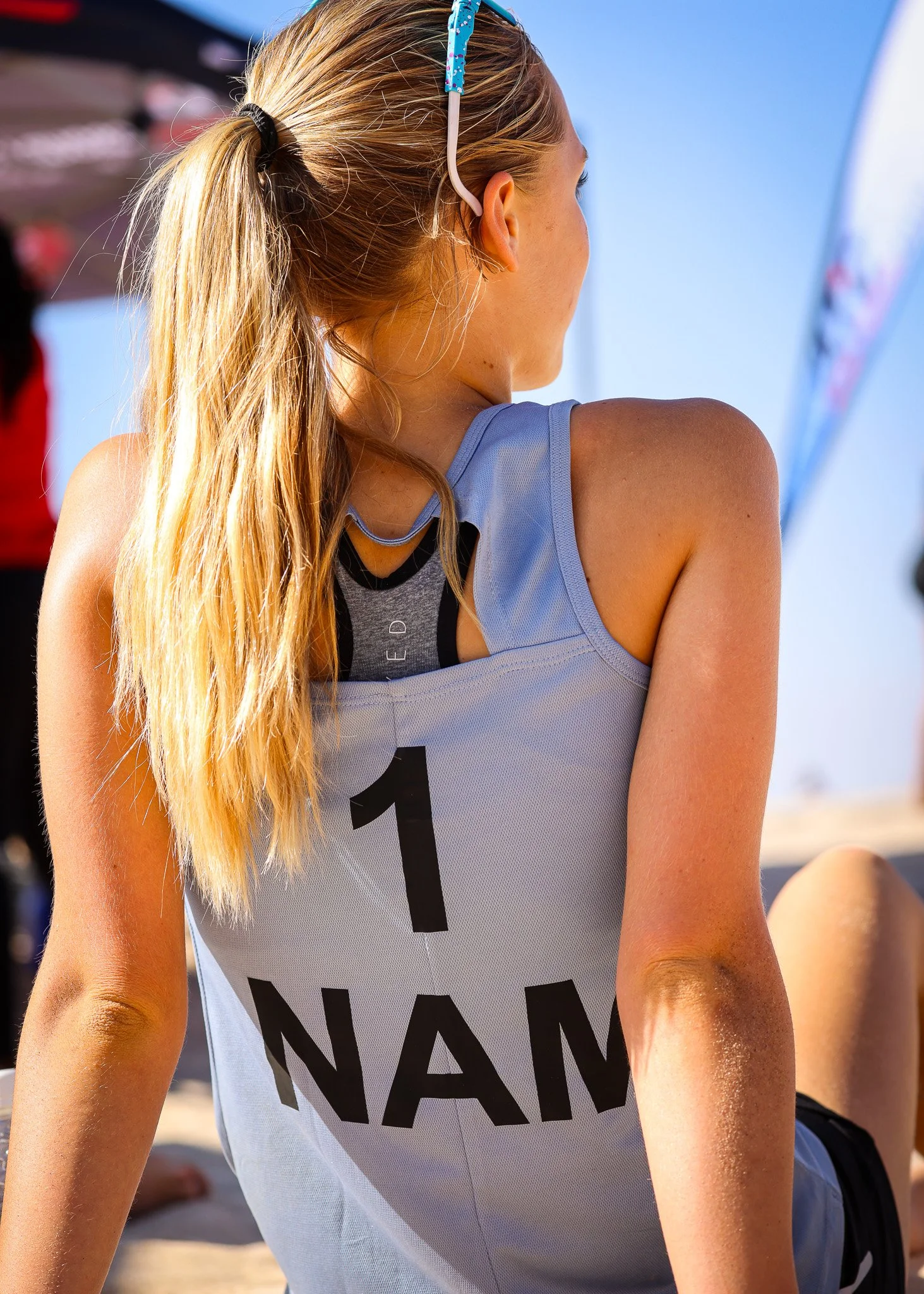 Back view of a female beach volleyball player with long blonde hair in a ponytail, wearing sunglasses on her head and a sports bikini top, sitting on the sand with a gray jersey labeled '1' and partially visible text.