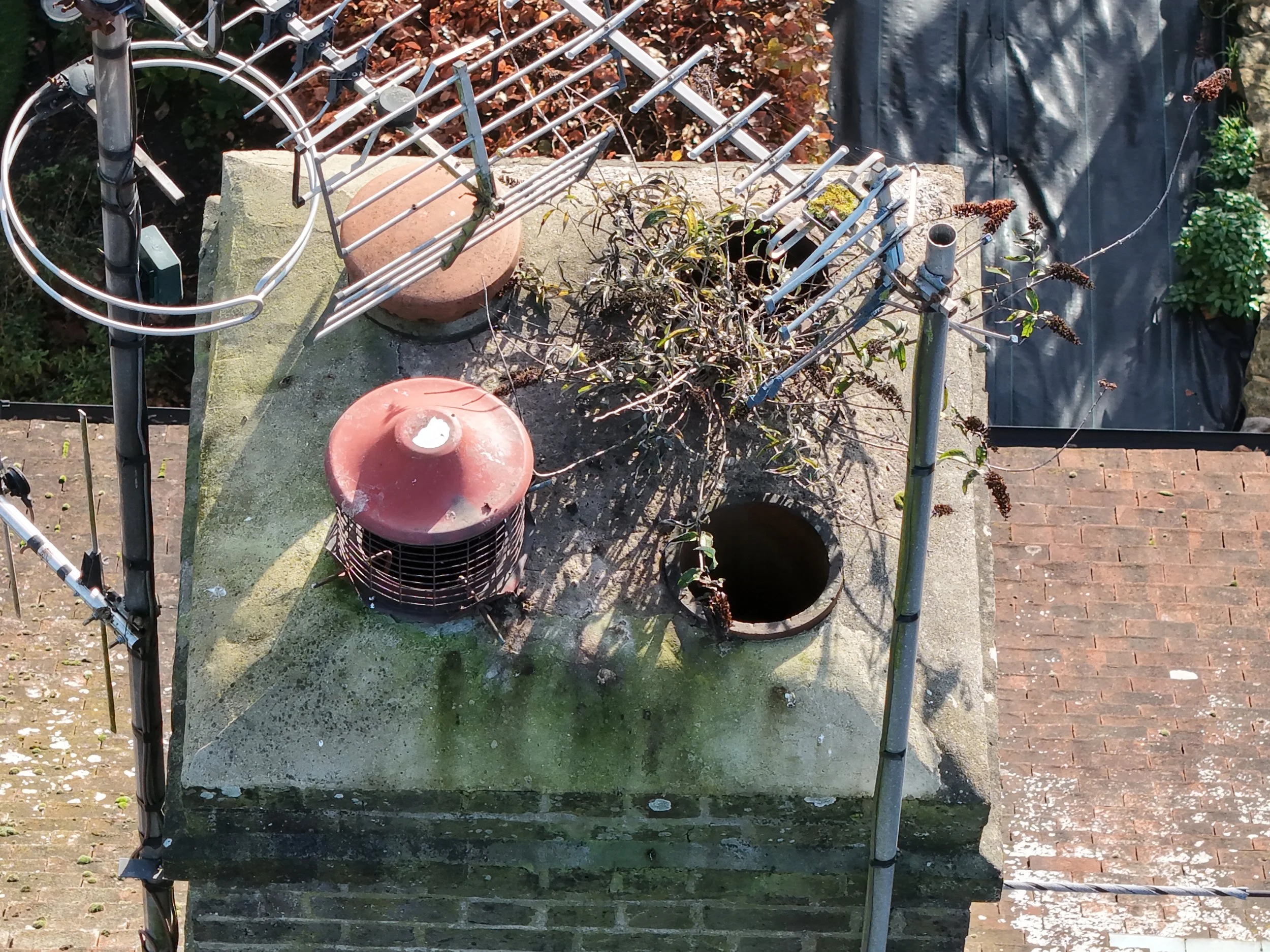 View of a brick chimney with an overgrown vine, an old dome-shaped vent, and TV antennas on top of the chimney. There is also a pipe opening and some black plastic sheeting covering an area nearby captured during a property drone survey inspection