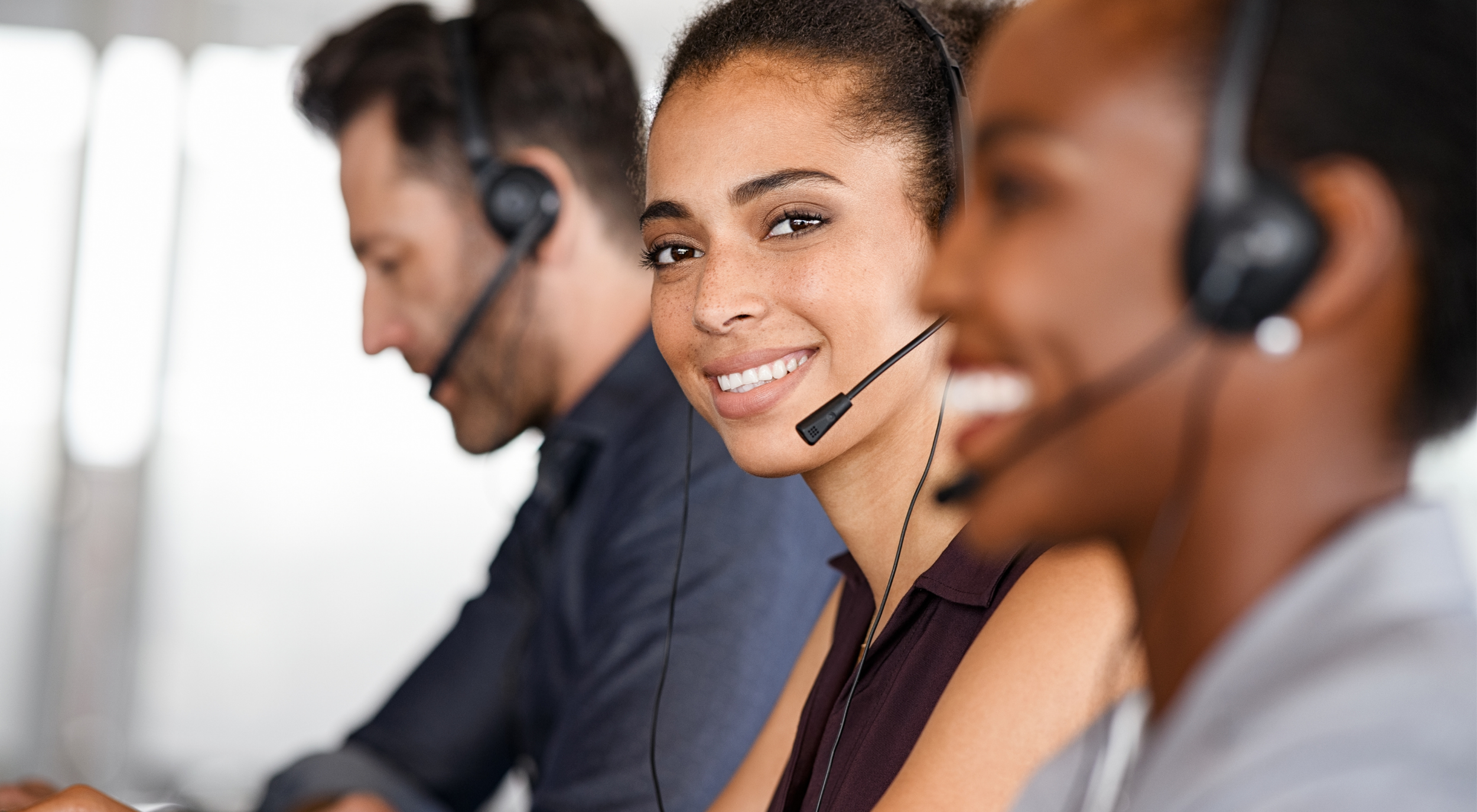 Tres personas en un centro de atención telefónica, usando auriculares con micrófono, una mujer en el centro sonriendo
