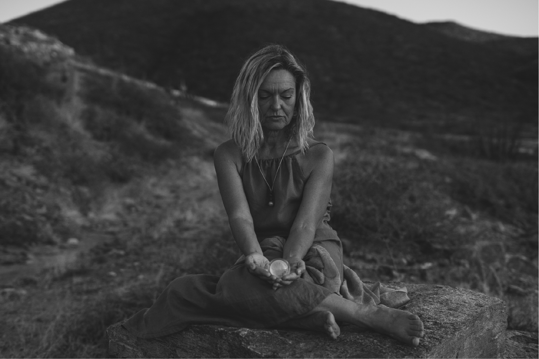 A woman with shoulder-length hair sitting cross-legged on a stone, holding a small round object in her open palms, outdoors with hills in the background, in black and white.