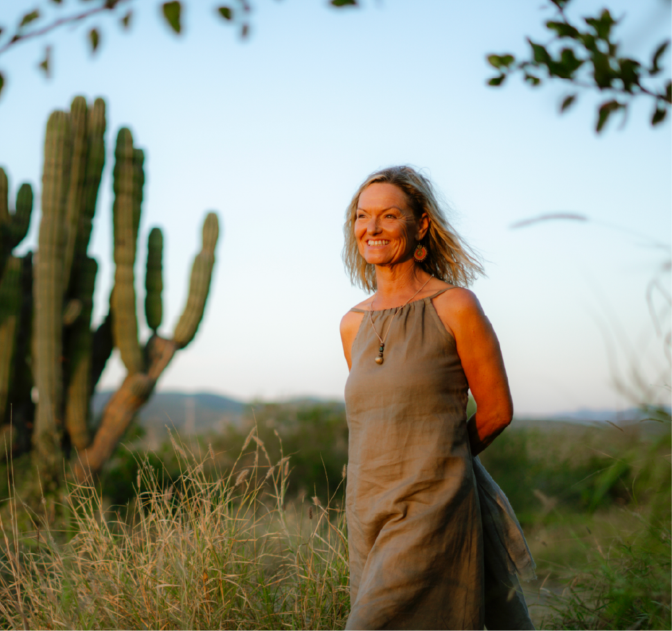 A woman smiling outdoors in a desert landscape with tall grass, a cactus, and a clear sky.