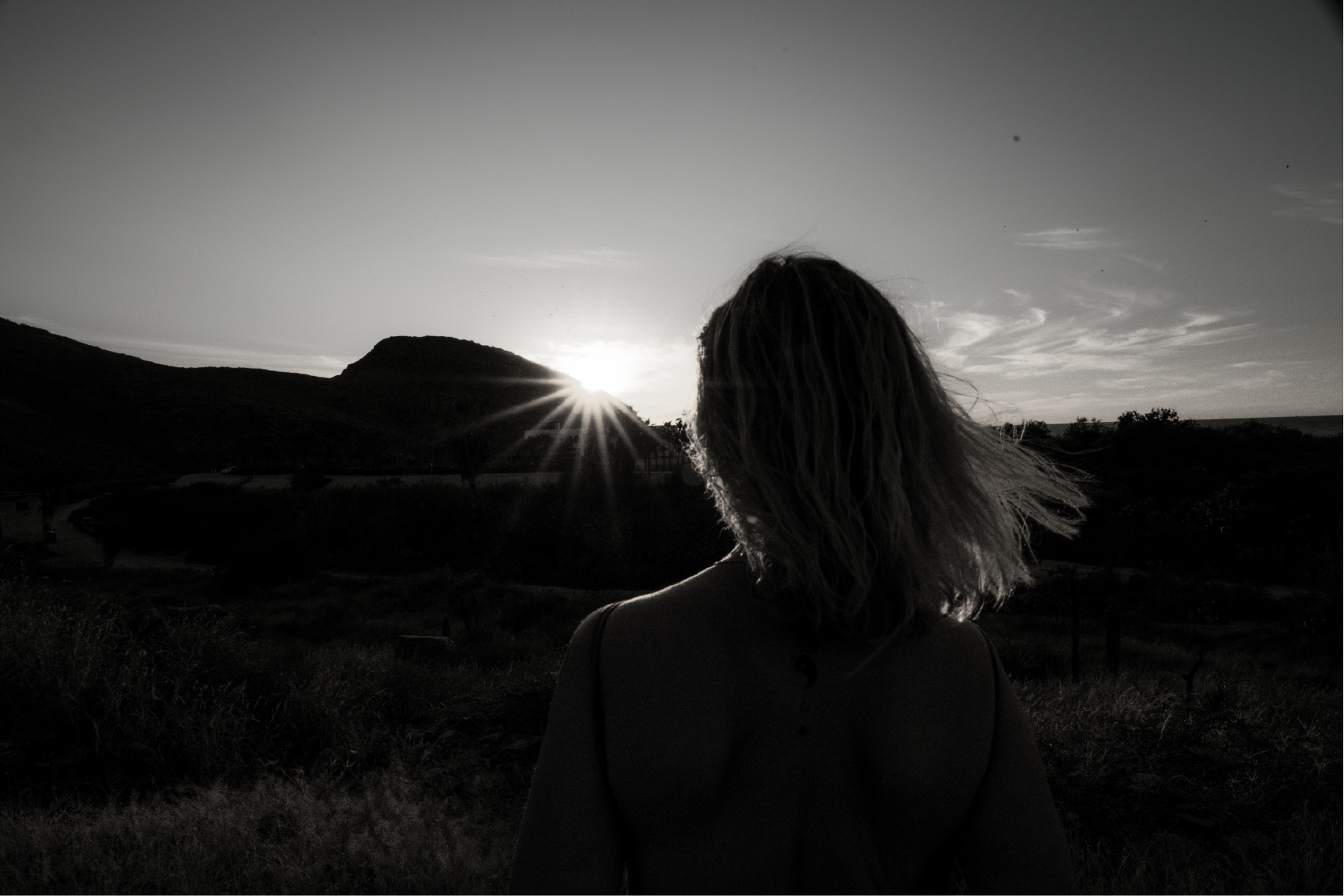 A person with shoulder-length hair seen from behind standing outdoors during sunset with hills in the background.